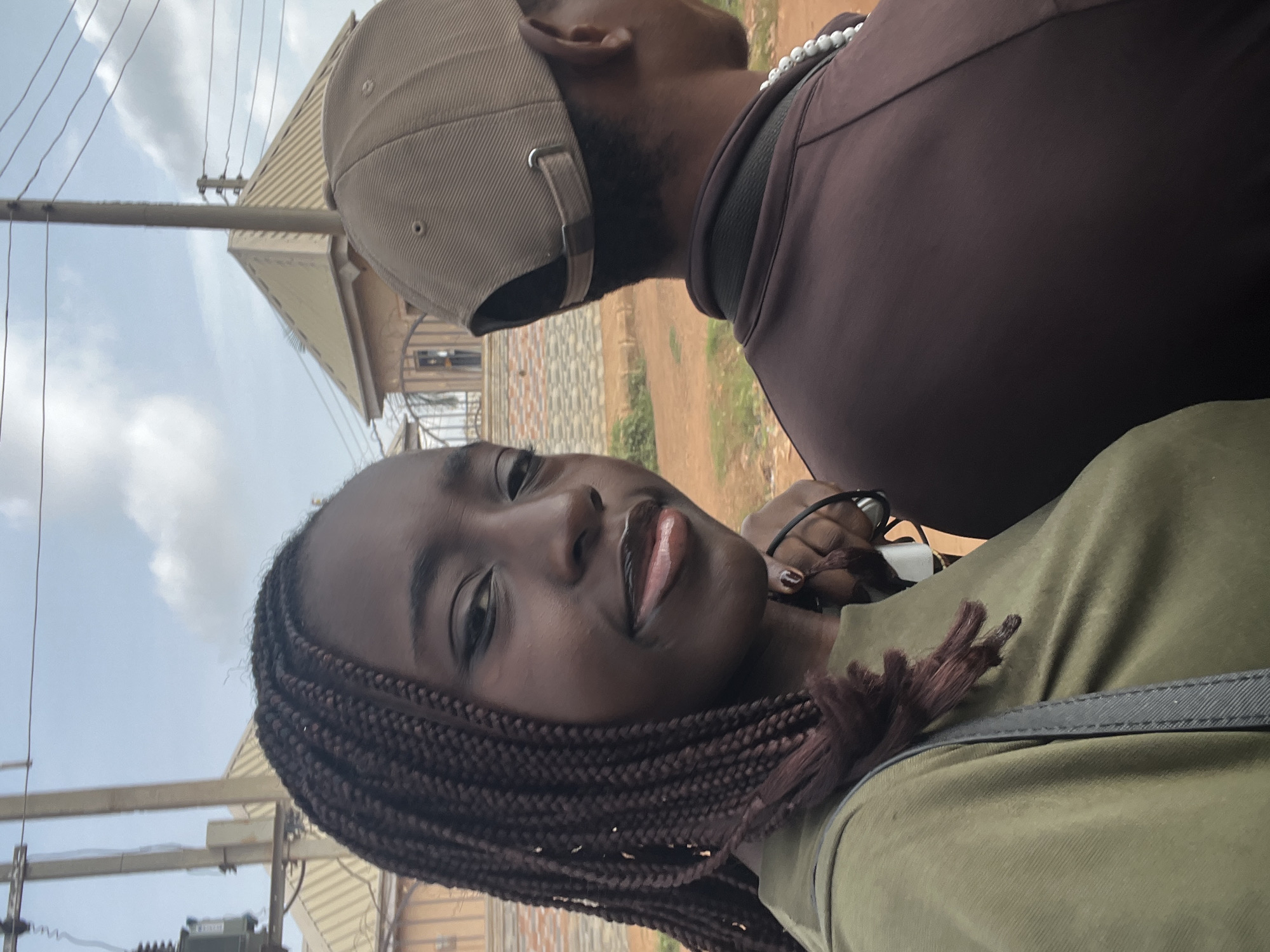 The image displays a close-up, outdoor scene featuring two individuals under a bright, cloudy sky. In the foreground, a young woman with dark skin and long, reddish-brown braided hair is looking directly at the camera with a subtle smile. She is wearing a light green top, and her lips appear glossy. Her right hand is visible near her ear, possibly holding a phone or earphones.
Behind her, slightly out of focus, is a man seen from the back of his head and shoulders. He is wearing a beige baseball cap, a dark brown long-sleeved top, and a white beaded necklace.
The background shows an urban or semi-urban environment. Utility poles with multiple electrical wires crisscross the sky, and an electrical transformer box is visible on one of the poles. Various buildings are present, including one with a light-colored, possibly corrugated roof, and another with a stone-patterned facade. A dirt road or path can also be seen in the mid-ground. The lighting suggests it is daytime. The scene evokes a casual moment in an everyday setting, fitting the context of Abuja, Nigeria. There is no visible text in the image.