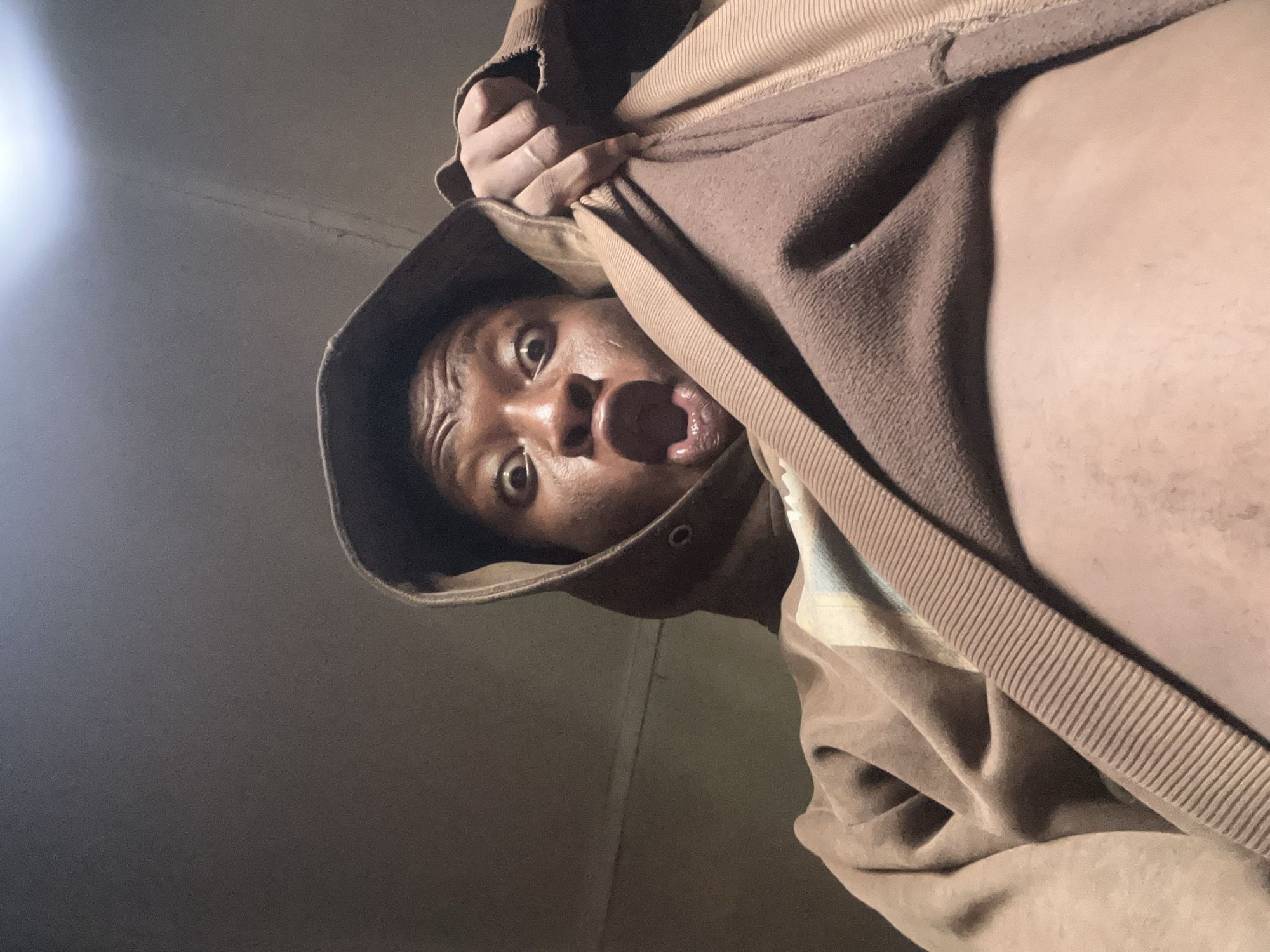 A person is holding up a brown hooded sweatshirt in a low-angle shot. The person is wearing the sweatshirt and is looking down at the camera with a surprised expression. The person's mouth is open and their eyes are wide. The background is dark.