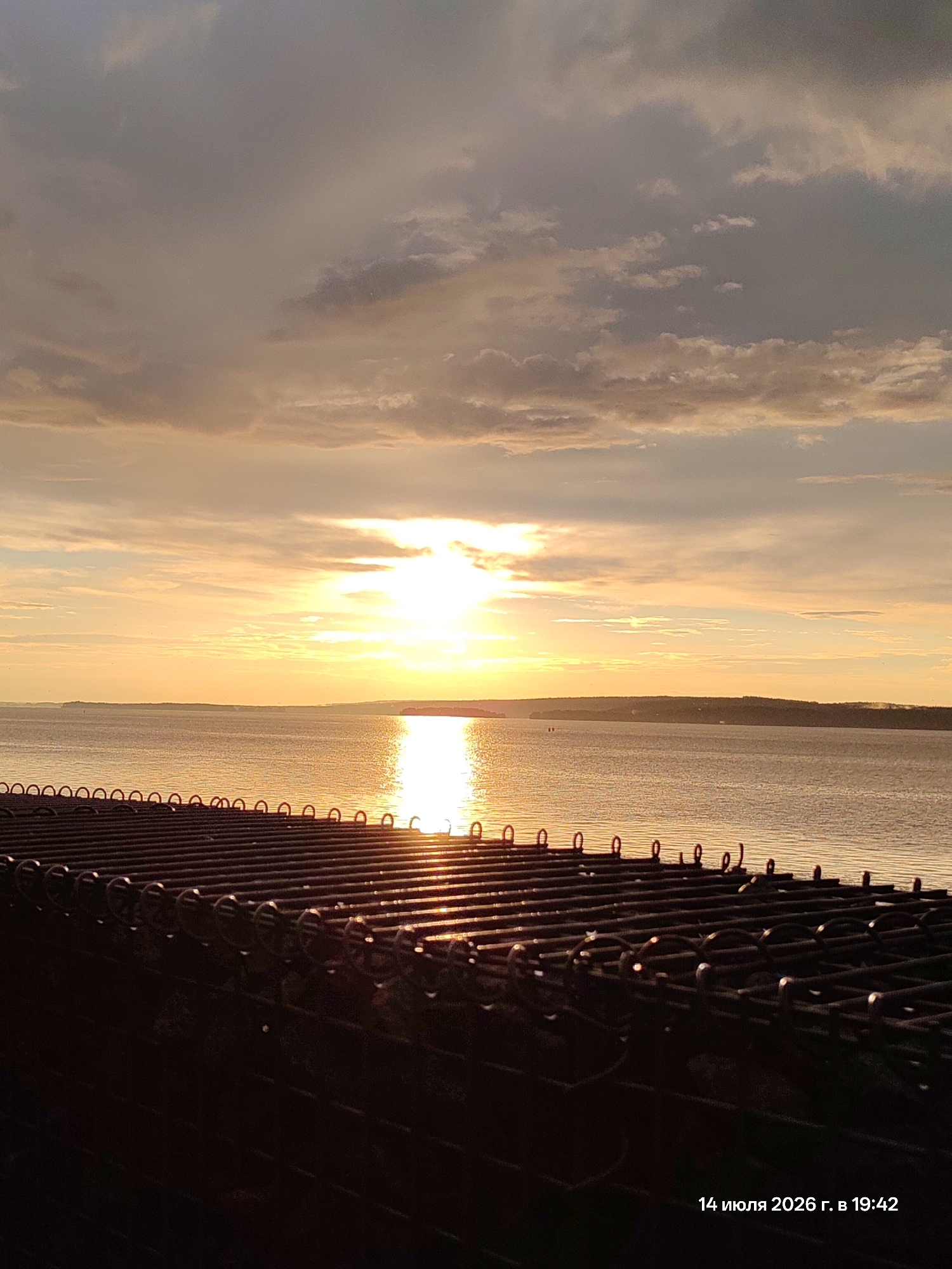 An outdoor, low-angle shot captures a sunset over a body of water, with a dark, metallic structure in the foreground and a distant shoreline under a cloudy sky. The sun, a bright, white orb, is partially obscured by clouds and casts a golden-orange glow across the water and sky, creating a strong reflection. The foreground consists of a dark, textured surface made of interconnected metallic rings and bars, arranged in parallel rows. The water is calm, with ripples catching the light. The distant shoreline is lined with a dark, indistinct band of land, dotted with faint silhouettes of trees or buildings. The sky is filled with a mix of dark gray and lighter, cloud-streaked hues, with the brightest part of the sunset concentrated in the center of the frame. A timestamp in the lower right corner reads "14 июля 2026 г. в 19:42".