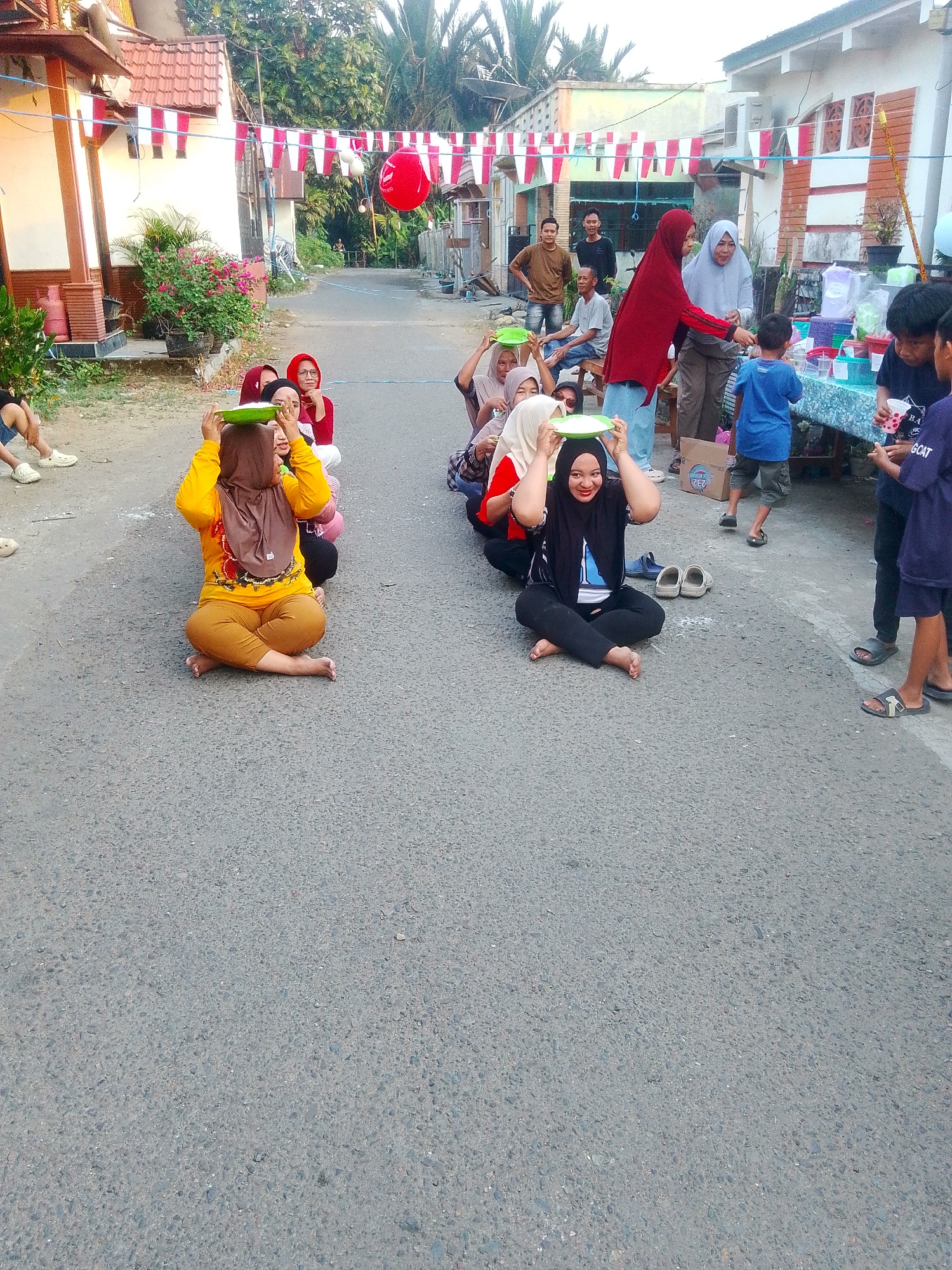 A group of people are participating in a game in a street in Barabai, Indonesia. In the foreground, several women are seated on the asphalt, balancing green plates on their heads. They are dressed in Islamic attire, including hijabs. Behind them, more people are standing and observing. A red balloon is visible in the upper portion of the image, attached to a string. Red and white decorative banners are strung across the street, suggesting a festive occasion. The environment appears to be a residential street with houses and trees.