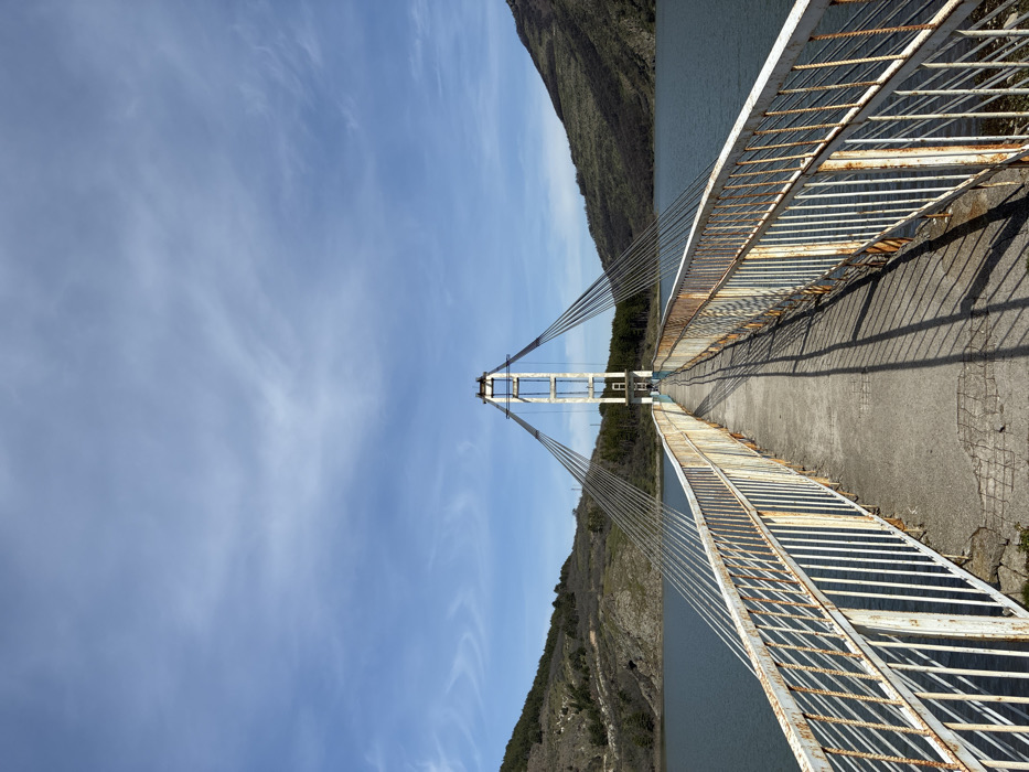 An exterior, eye-level shot captures a pedestrian cable-stayed bridge spanning a body of water, located in Lisitsite, Bulgaria. The bridge features a narrow, gray concrete walkway, displaying some cracks and wear. White-painted metal railings, heavily rusted with prominent orange and brown discoloration, line both sides of the walkway. Long, distinct diagonal shadows of these railings are cast across the concrete deck, originating from the right. A central white metal pylon, composed of two vertical supports connected by horizontal beams, rises from the mid-span of the bridge. Multiple metallic-gray suspension cables extend from the top of this pylon downwards to anchor points along the bridge deck's sides. The bridge crosses a calm, dark blue-green body of water, likely a reservoir. On both the left and right sides of the water, distant hills rise, covered with sparse green and brown vegetation, suggesting a dry or early spring landscape. The sky above is clear blue with scattered, wispy white clouds. No people or animals are visible on the bridge or in the surrounding environment. The scene is illuminated by direct daylight.