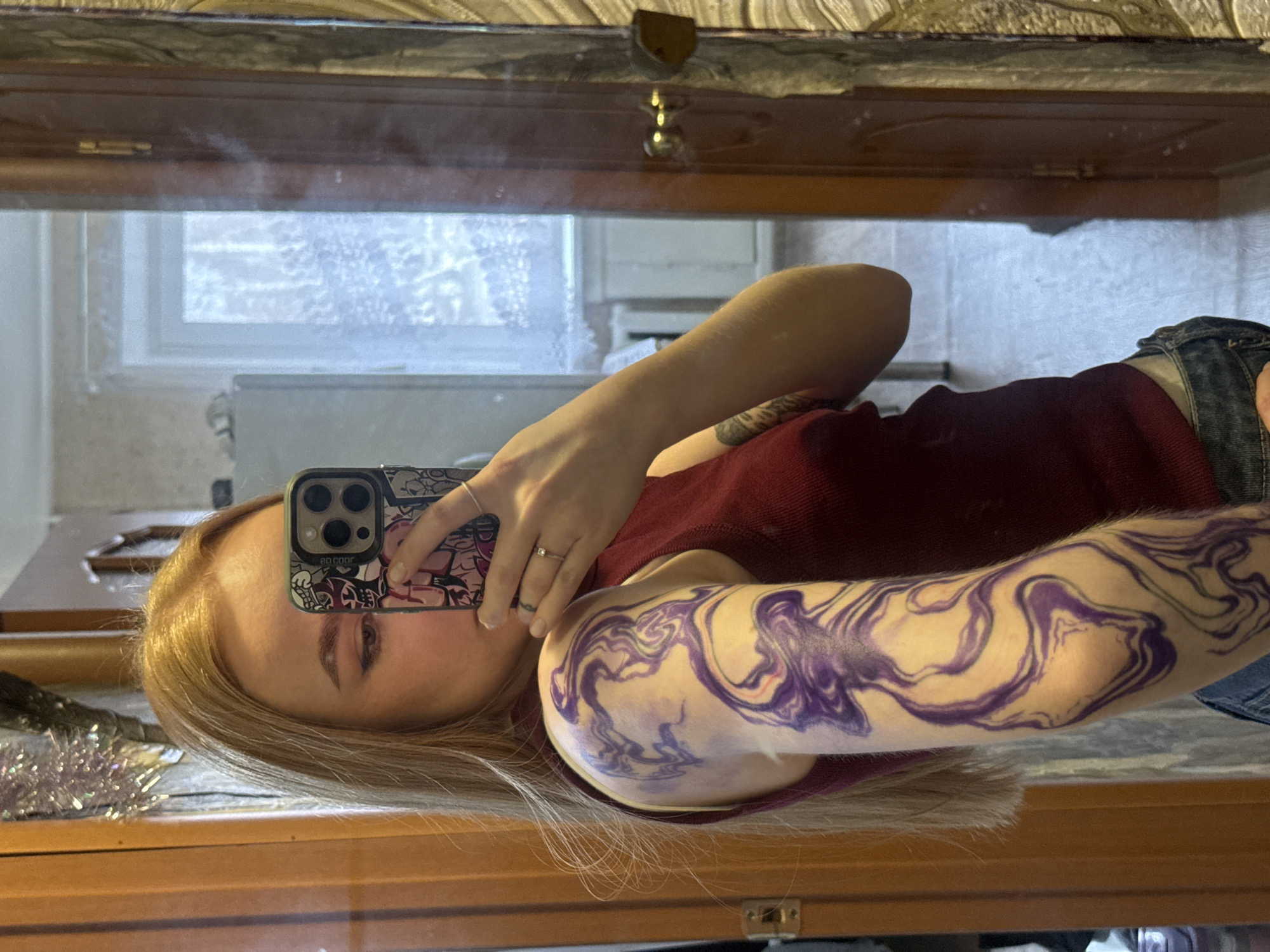 A woman with blond hair is taking a selfie in a mirror. She is wearing a red shirt and has a large, purple, marbled tattoo on her right arm. She is holding a smartphone with a patterned case to cover her face. The background includes a window with frost on the glass and a wooden cabinet. There is no information to confirm the location in the city of Chaikovsky, Russia.