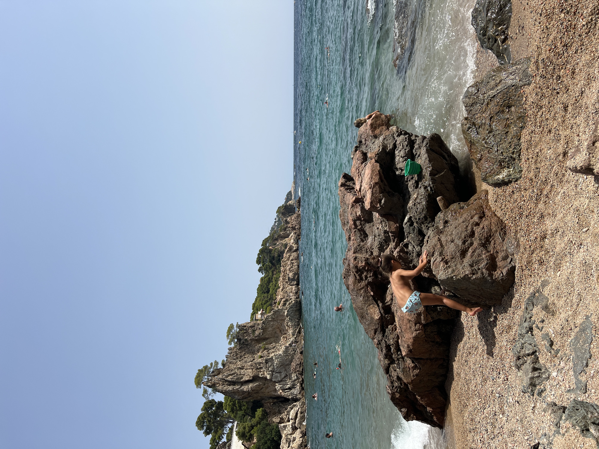 The image depicts a rocky beach on a sunny day, likely in Palafrugell, Spain, given the distinctive Mediterranean coastline. In the foreground, a young child with brown hair pulled back in a ponytail is on hands and knees, climbing over large, reddish-brown rocks on the sandy beach. The child is wearing patterned swim trunks. Behind the child, the sea is a vibrant turquoise color, with gentle waves lapping at the shore. Several people are swimming and enjoying the water in the middle ground. Further in the distance, a dramatic rocky outcrop covered in green vegetation rises from the sea. The sky is clear and pale blue. A small green bucket rests on a rock near the water's edge. The overall scene is one of a serene coastal vacation.