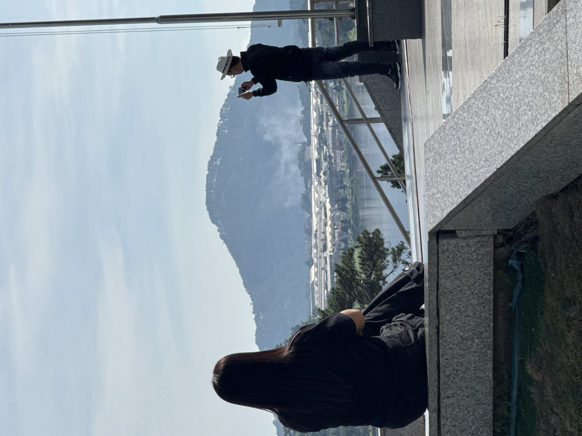 The image depicts a scenic viewpoint in Da Lat, Vietnam, overlooking a valley with a lake and distant mountains. In the foreground, a person with long dark hair is seated on a granite ledge, facing away from the camera and towards the vista. They are wearing a black top and dark pants. To the right, another person, a man wearing a white fedora, black jacket, and dark jeans, stands on a set of stairs and is taking a photograph with his mobile phone. A flagpole stands to the right of the man. The environment is characterized by clear skies, lush green vegetation on the slopes of the mountains, and structures in the valley below, including what appear to be greenhouses or industrial buildings. The overall impression is one of a peaceful, expansive landscape being appreciated by visitors.