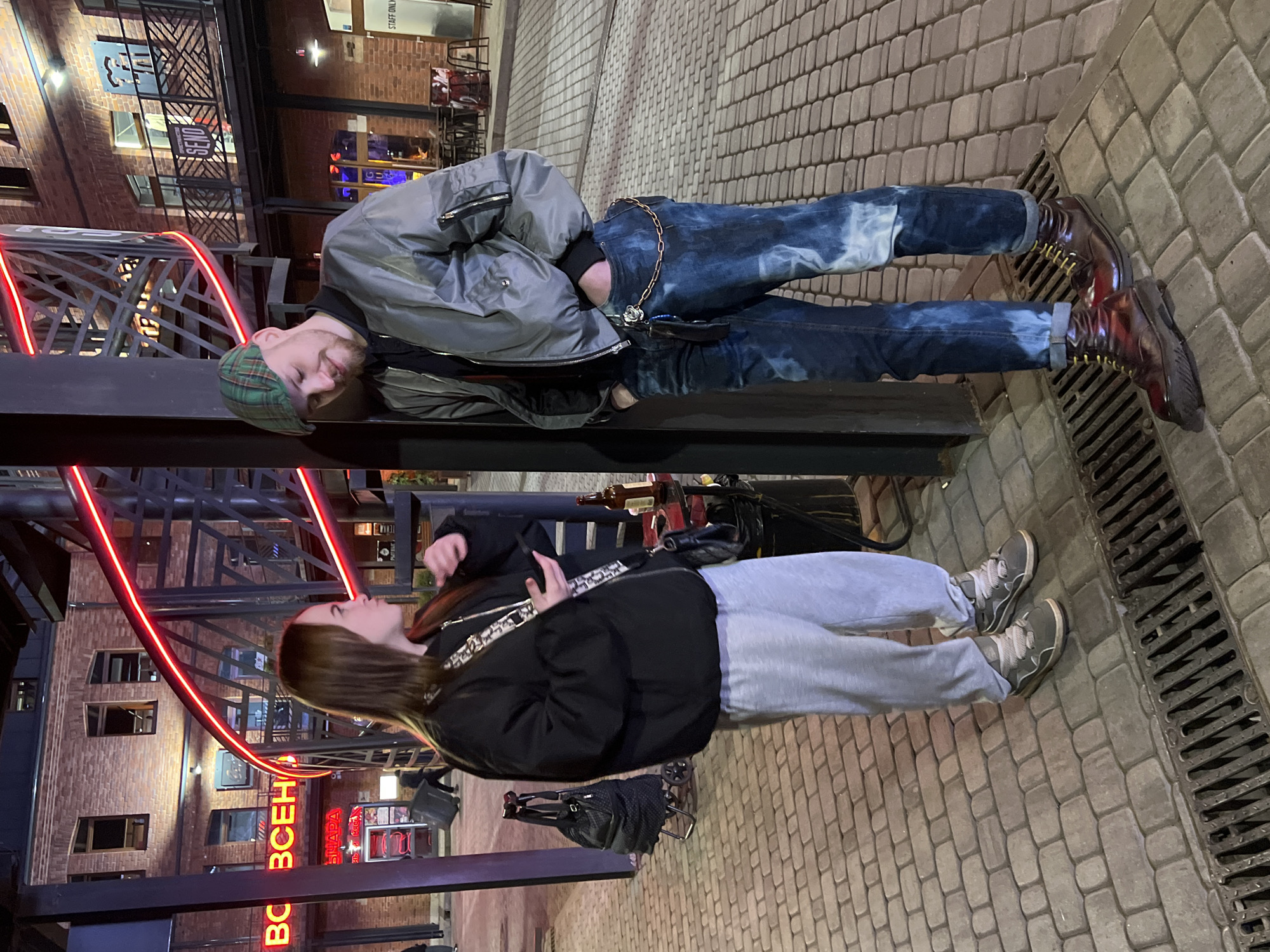 Two people are standing on a sidewalk in Saint Petersburg, Russia. One person is a male with a beard and green cap, wearing a grey jacket and blue jeans with chains. The other is a female with long brown hair, wearing a black jacket and grey sweatpants. They appear to be conversing. The background includes a brick building with red neon signs written in Cyrillic, and decorative metal structures with red neon lighting. The ground is paved with brick, and a metal grate is visible along the edge of the sidewalk.