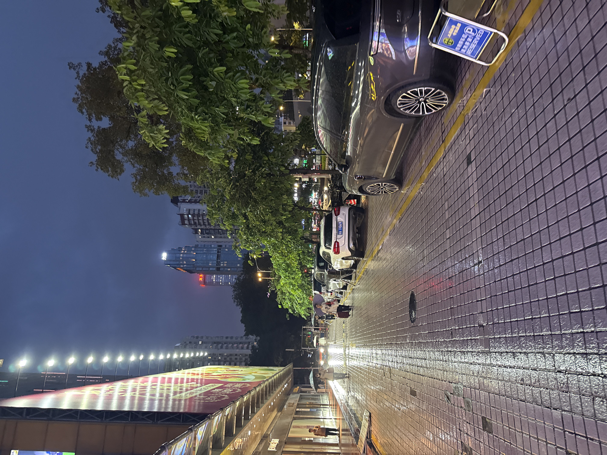 This is a sideways night-time shot of a street scene in Tianhe District, China. The sky is dark and appears to be overcast or twilight. The wet pavement reflects the streetlights, suggesting it might be raining or has recently rained.
On the left side of the image, there is a large building with a prominent advertisement banner that is brightly lit. The banner displays images and text in Chinese characters, though the text is not entirely legible at this resolution. Behind the banner, a row of streetlights stretches into the distance. Further back, modern high-rise buildings are visible, some with illuminated windows.
The right side of the image shows a sidewalk paved with small, dark tiles. Several cars are parked along the curb. One dark-colored car in the foreground has a blue and white sign attached to its rear bumper that reads "Reserved" in English and has Chinese characters as well. Next to it is a white SUV.
A line of trees with green foliage runs along the sidewalk, separating it from the street. People are visible walking on the sidewalk, some holding umbrellas, further indicating the possibility of rain. The overall atmosphere is that of a city at night, with the presence of rain adding a reflective and slightly somber mood. The lighting suggests it is dusk or early evening.