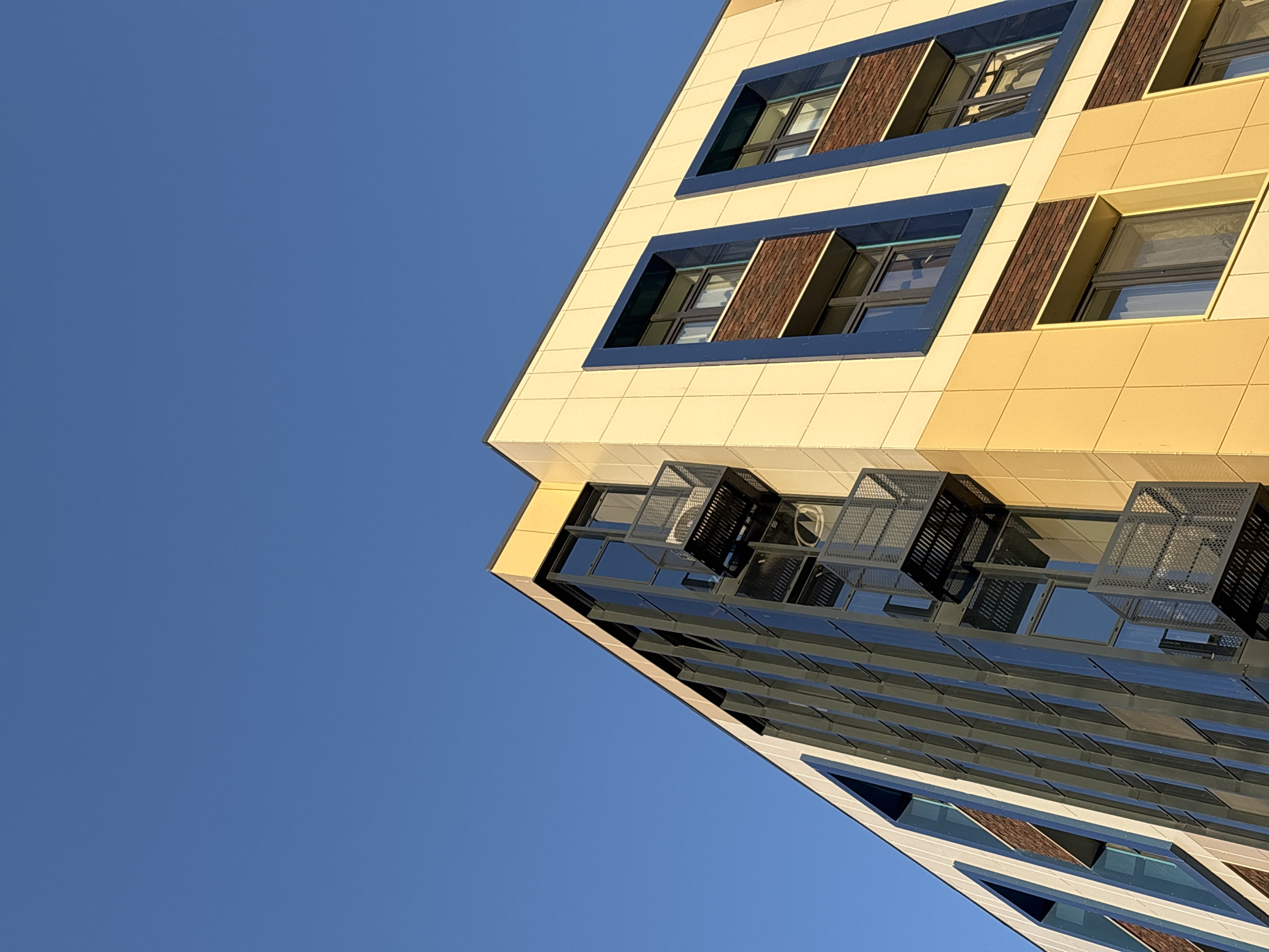 A low-angle shot presents the corner of a modern building against a clear blue sky. The building features a mix of architectural elements, including reflective windows and a cream-colored facade with rows of windows framed in dark blue and light brown. Ventilation units or air conditioning components are visible near the top section of windows. The building is located in Perm, Russia.