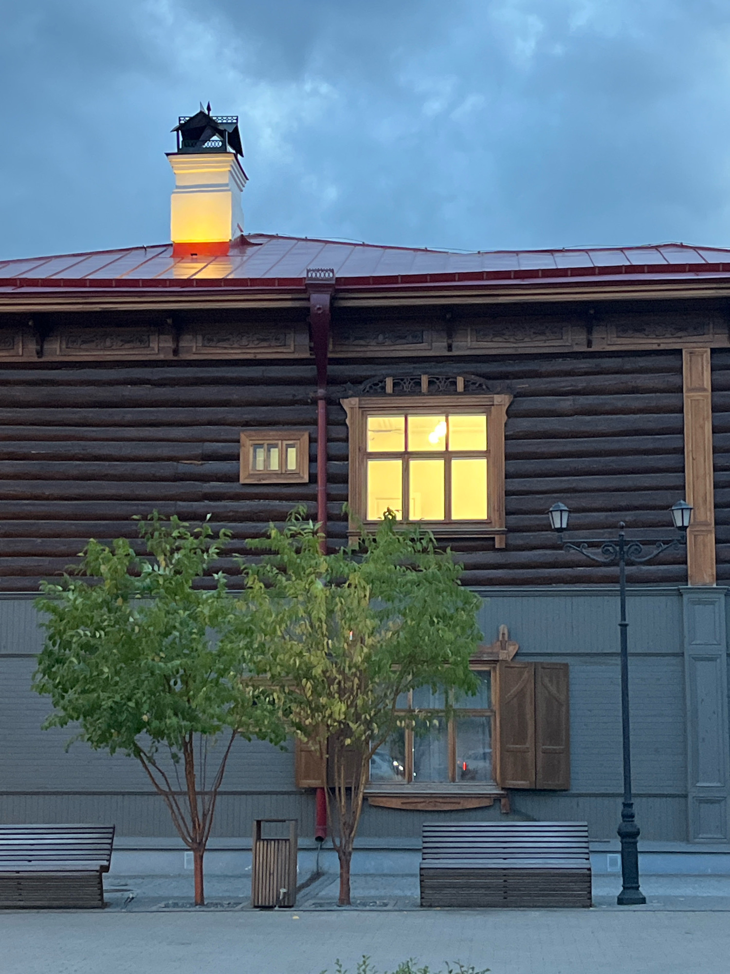 This image depicts a section of a traditional wooden building with a red metal roof and a decorative chimney. The building's exterior is constructed from dark, horizontal wooden logs, with ornate carved details above the windows and along the eaves. A large window on the upper floor is illuminated from within, casting a warm yellow glow. Below this, a smaller window with closed wooden shutters is visible. In front of the building, two young trees with green foliage stand beside a lamppost with two lights. Several wooden benches and a trash receptacle are placed on a paved area in front of the building. The sky is overcast with a twilight blue hue. This scene is located at the Strelka (Strelka, meaning "arrow" or "spit" in Russian) embankment in Krasnoyarsk, Russia, a prominent public space known for its historical architecture and views of the Yenisei River.