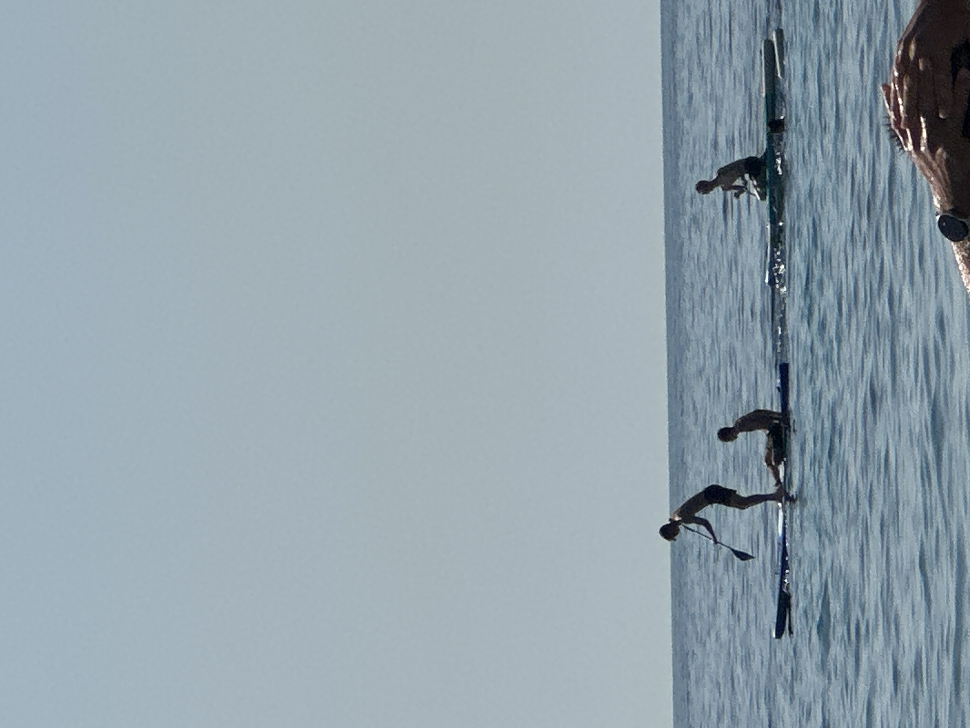 In the waters off Đurovići, Montenegro, three individuals are engaged in water activities. Two people are on paddleboards, one standing and paddling, the other seated. A third person is partially visible in the water, possibly having fallen from a board. The water is calm with gentle ripples, reflecting the clear, pale sky. The scene appears to be a peaceful daytime setting.