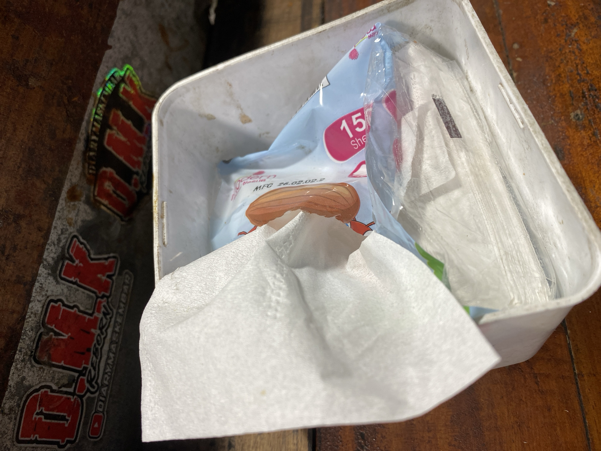 The image shows a white plastic bin filled with wet wipes. A white paper towel is draped over the edge of the bin. In the background, there are stickers with red lettering that say "D.M.K REBORN DIARYMASKEMBAD" and "D.M.K DIARYMASKEMBAD". The bin and wipes are placed on a wooden surface. The scene is indoors, possibly in a workshop or garage. The location is specified as Kasihan, Indonesia.