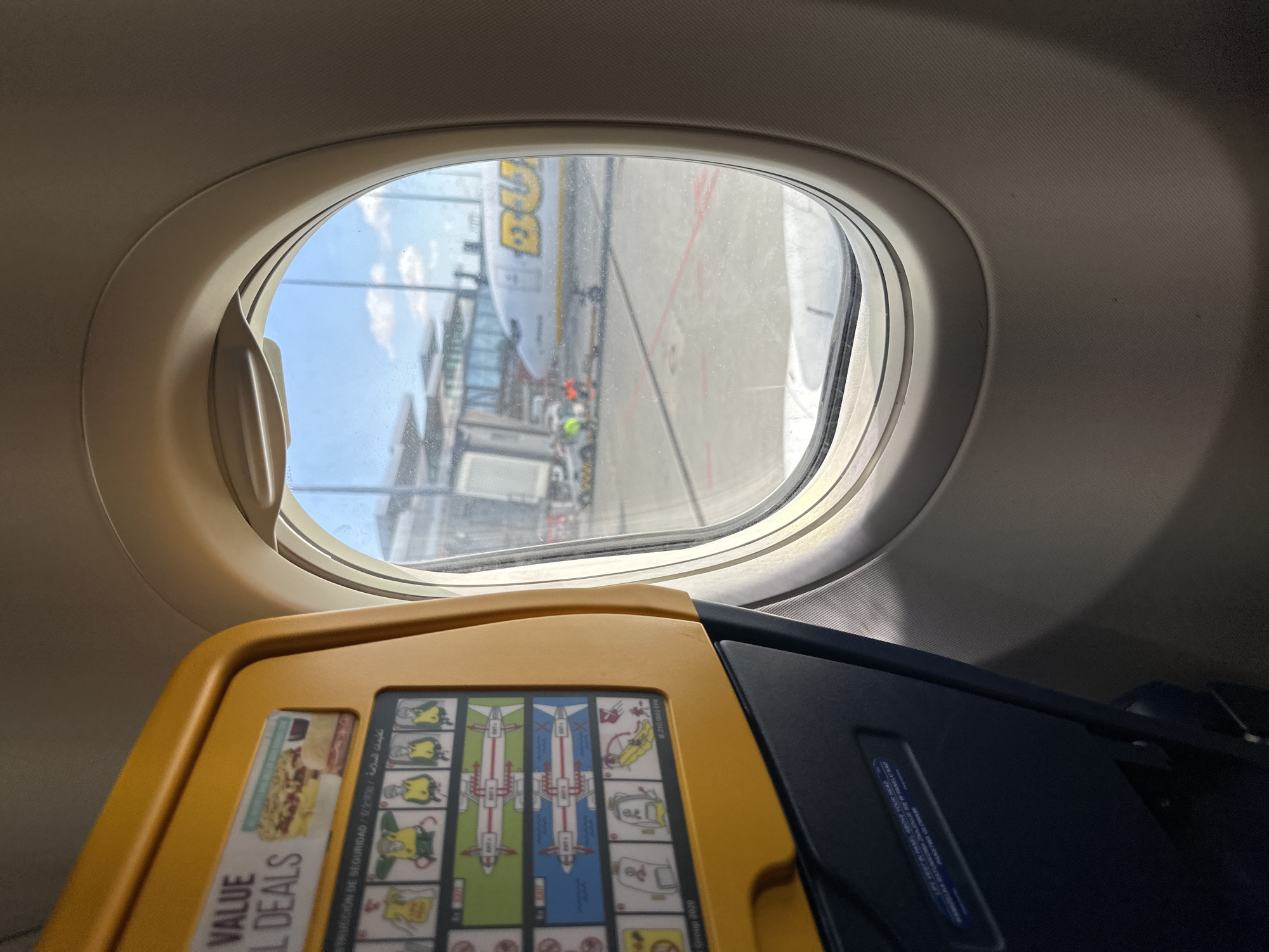 The image, taken from inside an airplane cabin, shows a view through a window of a Boeing 737-800, likely operated by Ryanair. The aircraft is parked at a terminal. Visible through the window is a section of the terminal building, some airport ground staff, and the wing of another aircraft. The foreground of the image shows the back of the seat in front, with a yellow tray table folded up, displaying an aircraft safety instruction card. The visible text on the safety card includes the phrases "VALUE DEALS," "INSTRUCCIÓN DE SEGURIDAD," "LIFE VEST IN PANEL ABOVE YOUR HEAD," and "KAMIZELKA RATUNKOWA ZNAJDUJE SIĘ W PANELU NAD." Based on the aircraft type, Ryanair's operation of this type, and the presence of Polish text on the safety card, it is inferred that the location is Wrocław Airport, Poland.
