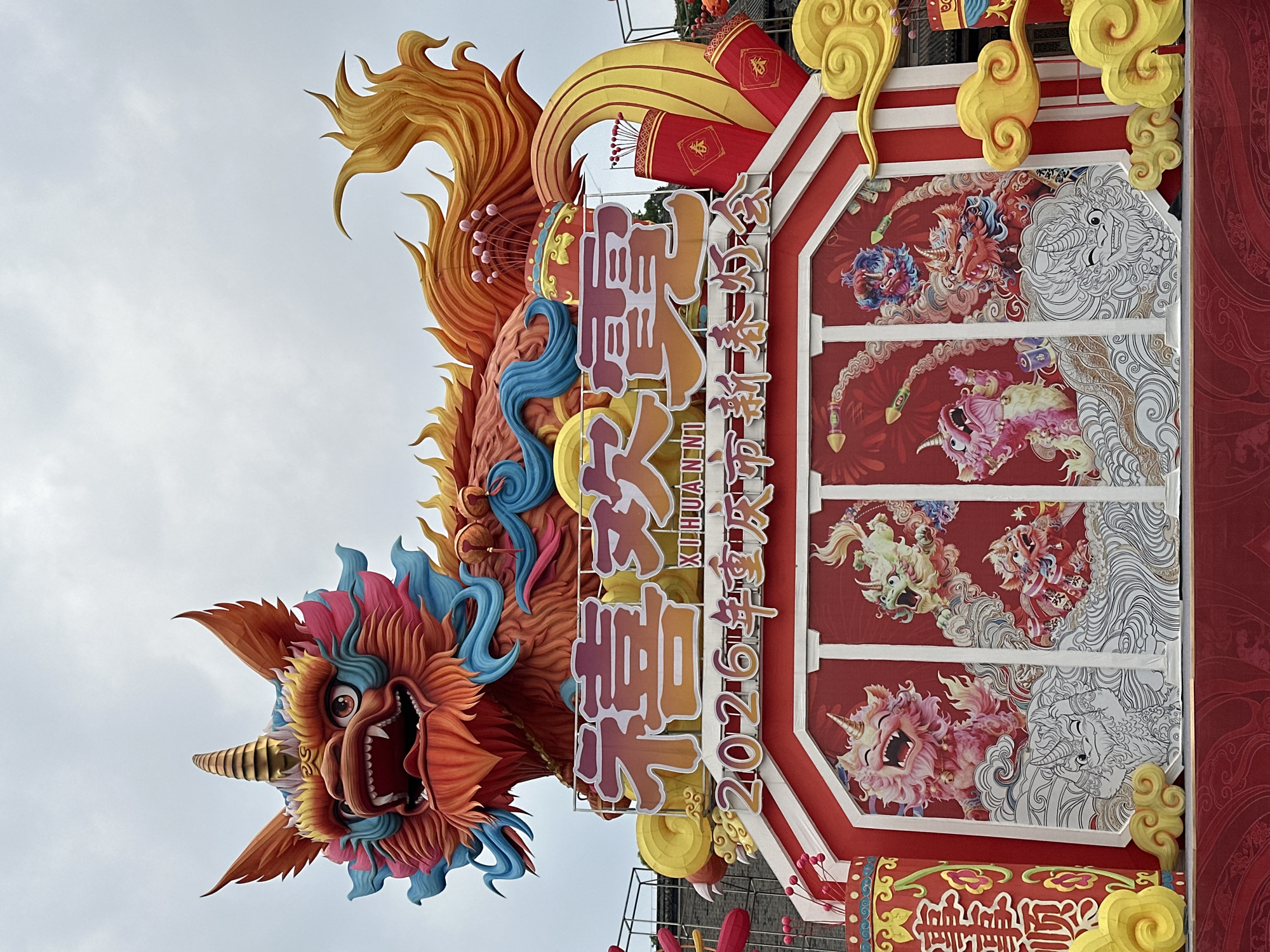 A large, colorful Chinese New Year decoration is the focus. It features a stylized dragon head with multi-colored scales and fur, a horn, and an open mouth, attached to a red and gold structure. The structure has Chinese characters and English lettering "XIHUANNI" (Xihuan Ni), along with "2026 年新年快乐" (2026 Nian Xin Nian Kuai Le, meaning "Happy New Year 2026"). The structure is adorned with images of various mythical creatures in vibrant colors. The background is a cloudy, overcast sky. The location is Liangjiang, China, as the characters translate to "Happy New Year 2026".
