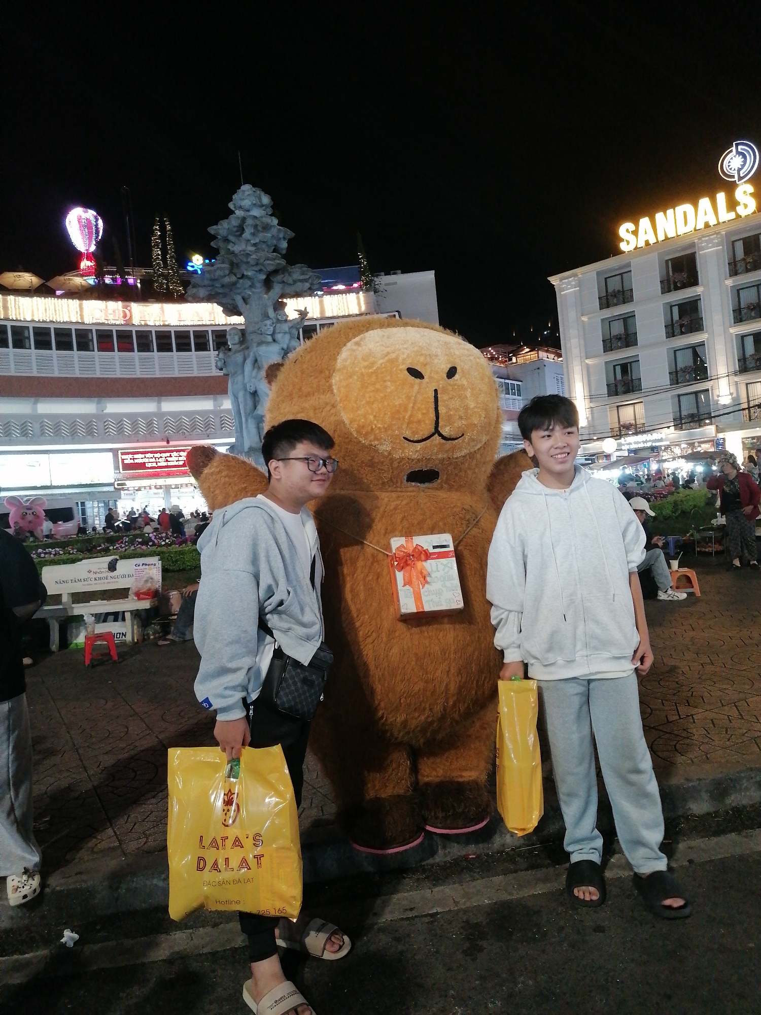 This is a vertical, full-shot photograph taken at night in Da Lat, Vietnam. The scene depicts a festive outdoor environment, likely a market or entertainment area. In the foreground, two young men are posing with a large, furry, brown bear mascot. Both individuals are wearing casual hooded sweatshirts and sweatpants, and they are holding bright yellow shopping bags. The man on the left is wearing glasses and has a dark crossbody bag. The man on the right is smiling broadly.
The bear mascot has a simple cartoonish face and is adorned with a red and white gift box tied with an orange ribbon, prominently displayed on its chest. Behind the subjects, a large white building with the illuminated neon sign "SANDALS" is visible at the top. Below this, there are other buildings with lights, suggesting commercial activity. To the left, a statue of people is partially visible, and further in the background, a lit-up hot air balloon can be seen against the dark night sky. Other people and stalls are scattered throughout the scene, indicating a lively atmosphere.
The yellow shopping bag held by the man on the left has text printed on it, which includes "LATA'S DALAT" and "ĐẶC SẢN ĐÀ LẠT" (Specialties of Da Lat), along with a hotline number. The overall impression is a cheerful, lively evening outing, possibly during a holiday or special event in Da Lat. The lighting is artificial, coming from various signs and structures, and the time of day is clearly night.