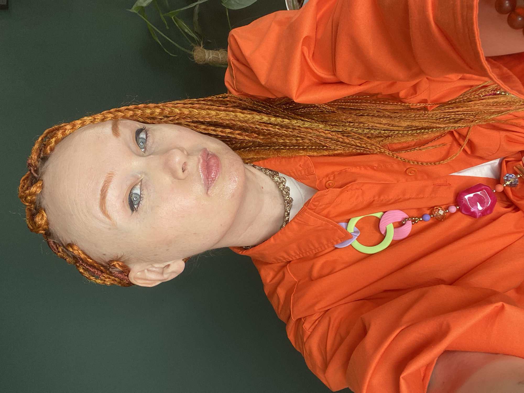 An overhead, tilted portrait captures a light-skinned individual looking directly at the camera. The person has light blue eyes, and their fair skin is subtly flushed. Their eyebrows are well-defined and a light brown color. Lips are parted slightly and adorned with a light pink gloss. Their hair is styled in numerous long, thin braids that are a vibrant orange-gold color, framing their face and extending down their shoulders.
The individual is wearing a bright orange collared shirt, with a hint of a white garment visible underneath. Around their neck, a delicate gold-colored chain necklace is visible, peeking out from under the collar. Over the orange shirt, a prominent and colorful necklace is worn, featuring a variety of large, playful beads and shapes including rings in light green, pink, and lavender, along with spherical beads in varying shades, and a large, glossy pink heart-shaped pendant. Another partial necklace with reddish-brown beads is visible on the right side of the frame.
The background consists of a solid, dark teal or deep green wall. In the upper left corner and partially in the upper right, the dark green leaves of a plant are visible, suggesting an indoor setting. The lighting is soft and even. No text is visible in the image. The image is presented as if taken in Sochi, Russia, though no specific visual cues confirm this location.