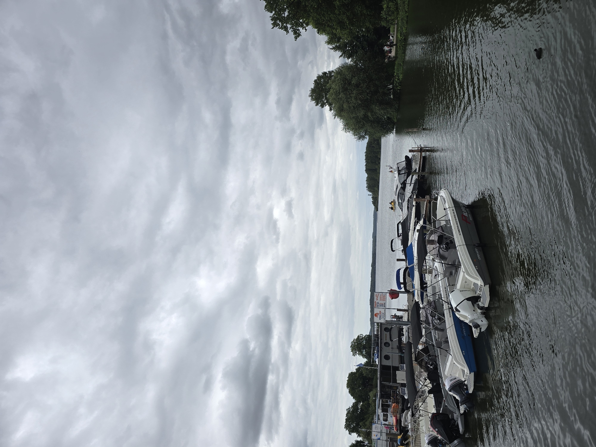 The image depicts a cloudy day at a marina in Schwerin, Germany, likely by the Pfaffenteich lake or the Ziegelsee lake, given the city's geography. In the foreground, several motorboats are docked at a wooden pier. These boats are of various sizes and designs, with some featuring outboard motors. The water is calm with small ripples. Behind the boats, a large houseboat or a floating structure is moored. Further in the background, the shoreline is lined with lush green trees, and a few people can be seen walking or sitting near the water. The sky is overcast with heavy, gray clouds, suggesting a diffused light condition. The overall scene conveys a peaceful, albeit somewhat somber, atmosphere of a lakeside recreational area.