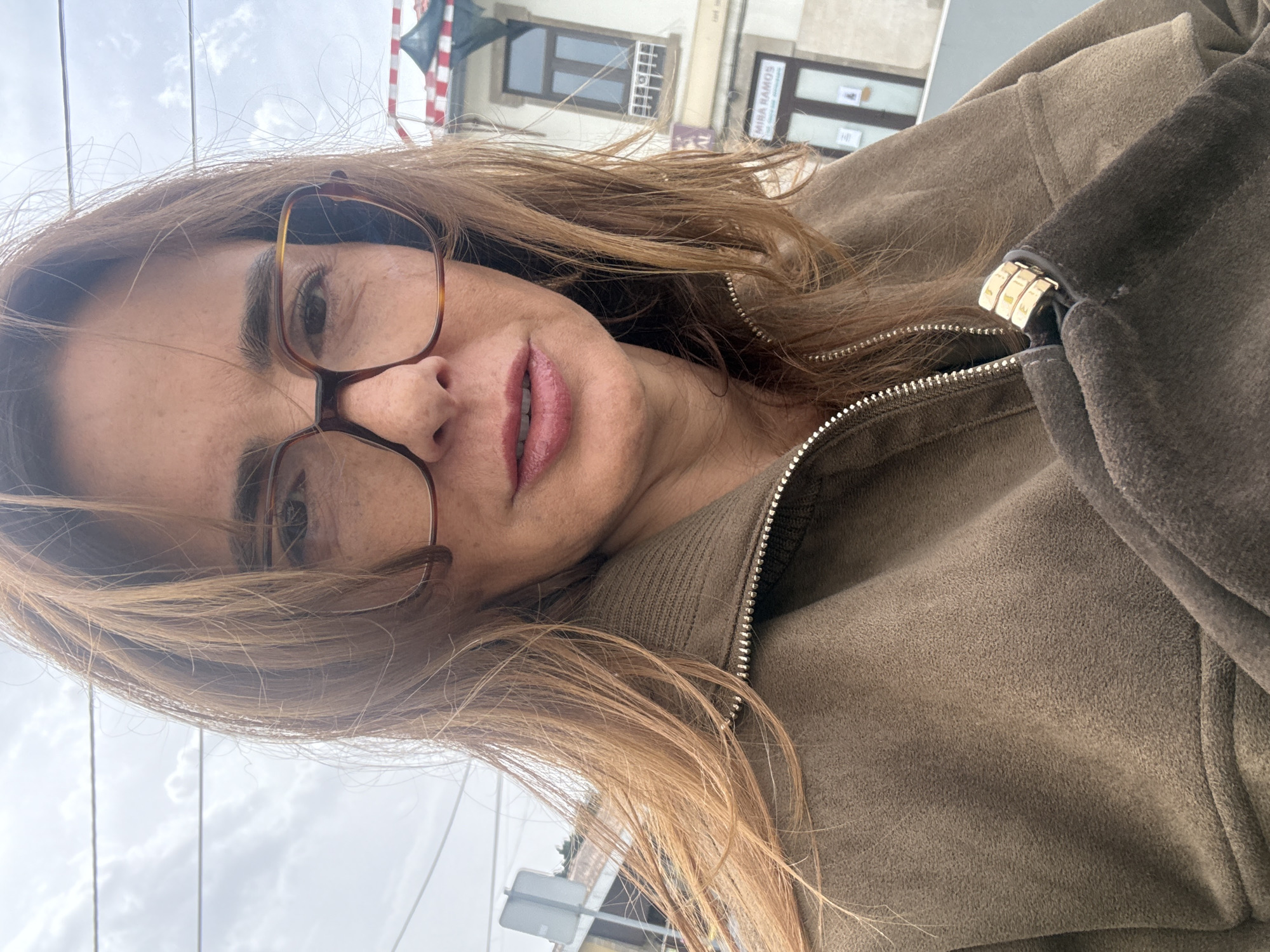 A close-up, rotated portrait shows a woman in an outdoor urban setting in Vila Nova de Gaia, Portugal. She has long, light brown hair and is wearing brown-framed glasses and pinkish-red lipstick. Her expression is calm and neutral as she looks towards the camera. She is dressed in a brown or olive-green jacket, likely made of a textured material such as suede or fleece, featuring a silver zipper and a gold-colored metallic buckle or clasp detail near her right shoulder.
The background reveals a cloudy sky and parts of buildings. One building has a red and white striped awning or banner, and another distinctly displays the text "MIRA RAMOS" on a sign, providing a clear location cue. The scene is illuminated by natural daylight, suggesting an overcast day. The image is composed with the woman's head tilted significantly to the left, indicating a diagonal capture or rotation.