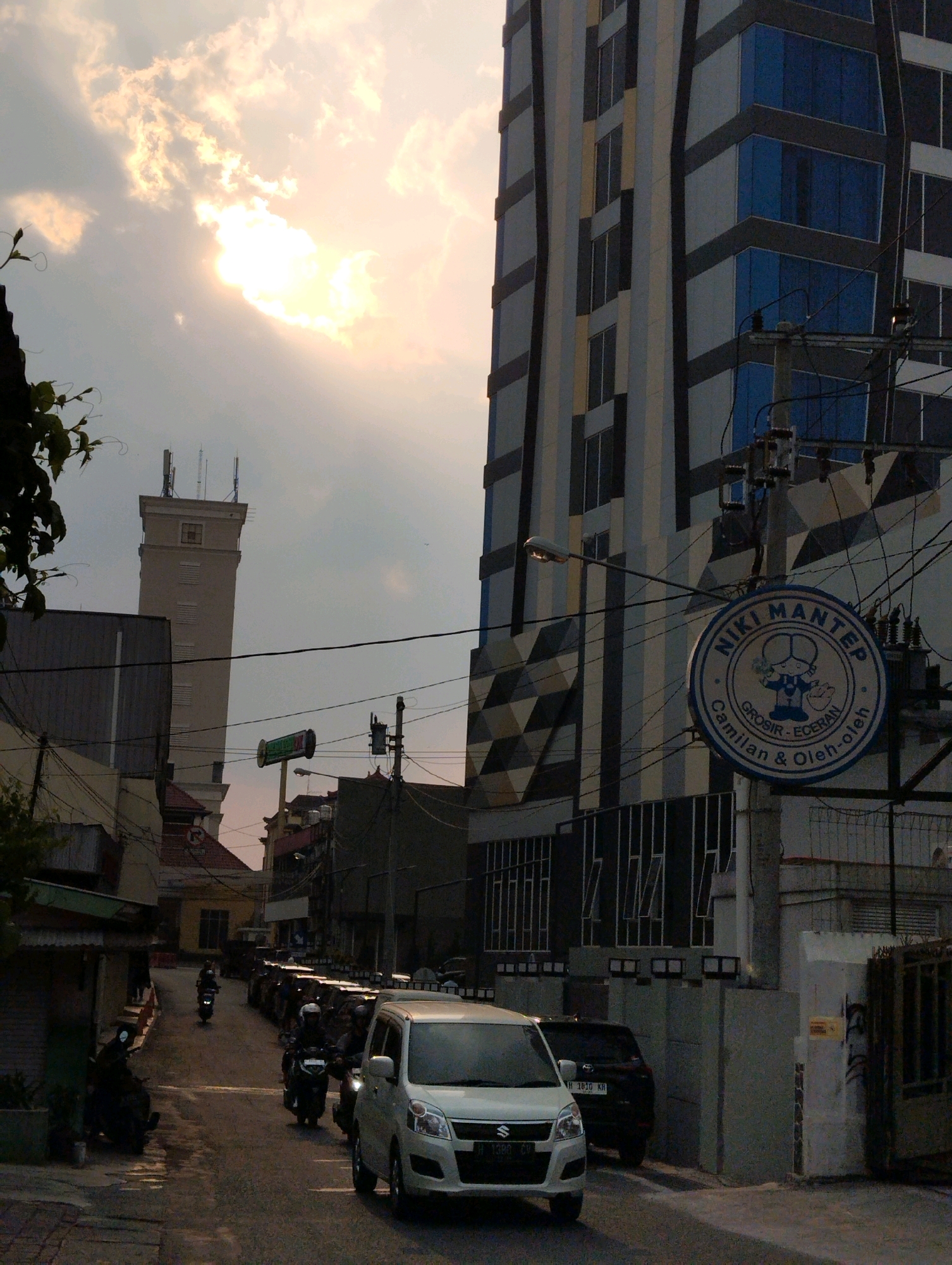 The image depicts a street scene in Salatiga, Indonesia, characterized by a mixture of modern architecture and older buildings. A tall, modern building with a facade of blue glass windows and geometric patterns dominates the background. To its left stands a distinctive, older tower structure with antennae on top. The street is lined with various buildings, some appearing to be commercial establishments, indicated by signage such as a circular blue and white sign with text that appears to read "NIKI MANTEP GROSIR - ECERAN Camilan & Oleh-oleh".
The street is populated by several motorbikes and a white car. Two individuals are visible riding motorbikes in the middle of the street, and another person is seated on a motorbike parked on the left side of the road. The sky is partly cloudy with the sun breaking through, casting a warm light and creating a slightly hazy atmosphere. The overall impression is that of a bustling urban environment during daytime.