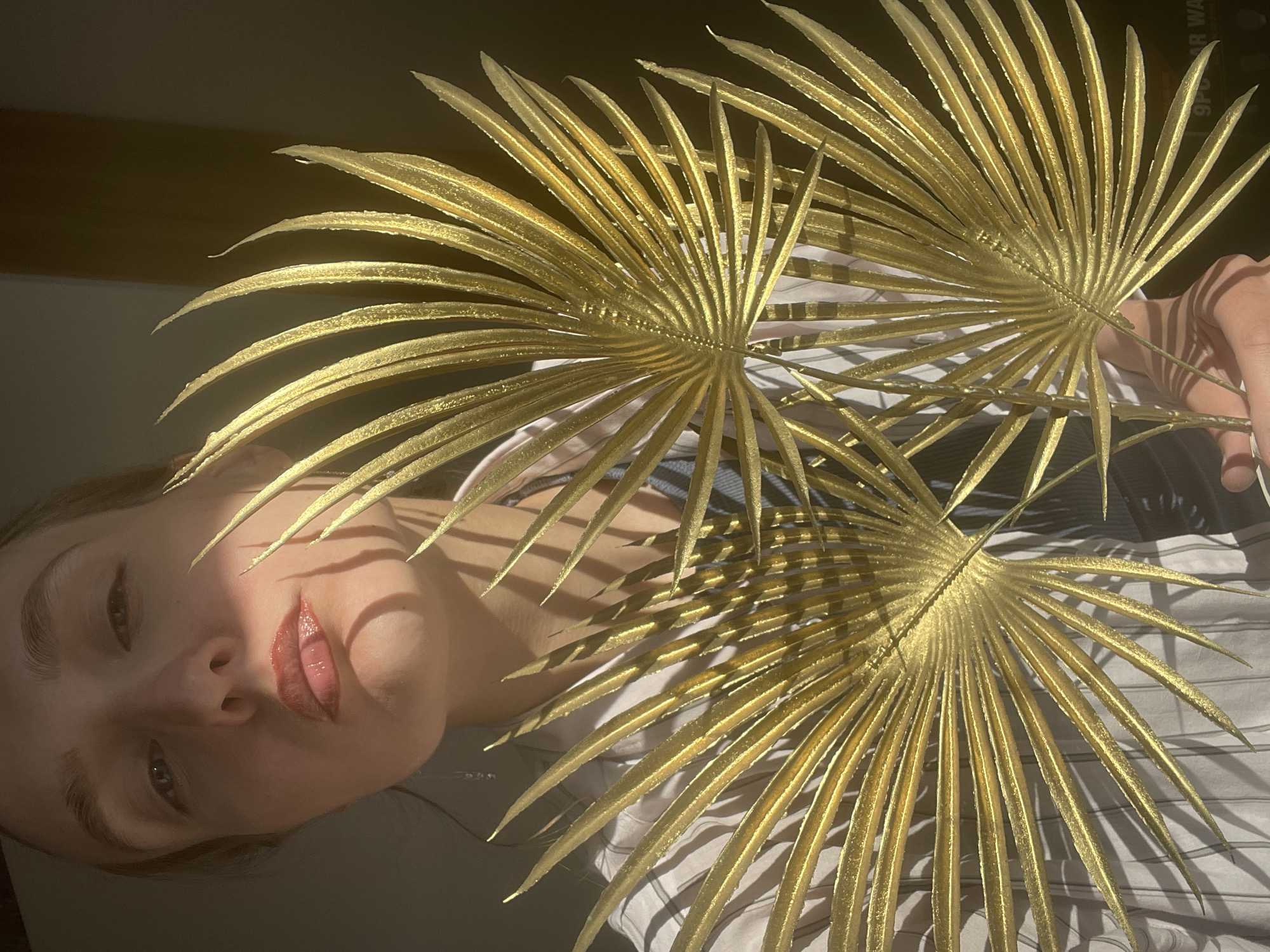 A selfie-style photograph depicts a person holding three gilded palm fronds in front of their face. The person, presumably female, has fair skin, visible eyebrows, and glossy lips. They are wearing a collared shirt with dark stripes underneath. The palm fronds cast shadows across their face. The background is out of focus and appears to be an interior space with a light-colored wall. The scene is in Sepych, Russia.