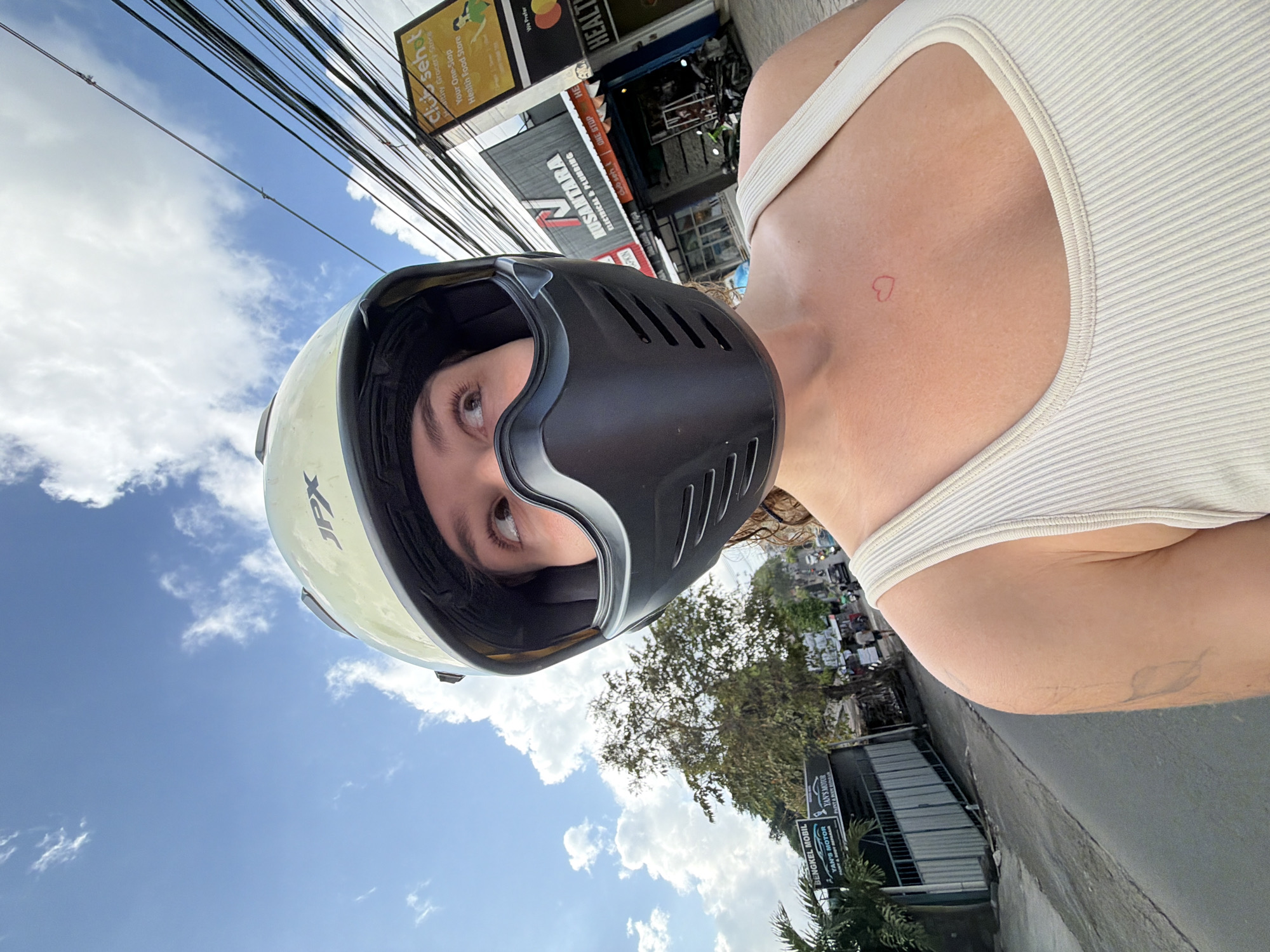 A medium shot captures a woman wearing a pale yellow helmet and a black face mask, looking upwards and to the right. She has curly brown hair and is wearing a white ribbed tank top. A small red heart tattoo is visible on her upper chest. The background shows a street scene in Mengwi, Indonesia, with buildings, power lines, and trees lining the road. One building has a sign that reads "NUSA NTARA ELECTRICAL & PLUMBING" and another sign says "Club Sehat Healthy Grocery Store". The sky is blue with scattered white clouds. The overall impression is of a person on the move, likely on a scooter or motorcycle, in an urban or semi-urban environment.