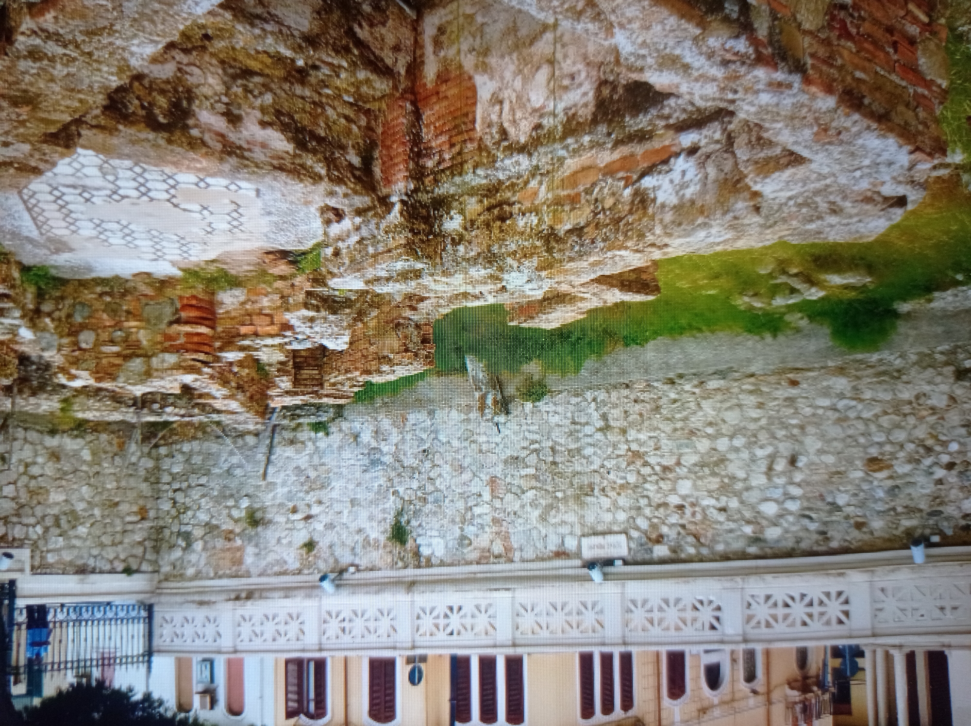 The image depicts an archaeological site with ancient ruins in the foreground, characterized by weathered brick and stone structures. The ruins are partially overgrown with grass and moss, suggesting a period of disuse. In the background, a stone wall supports a decorative white railing, beyond which a building with arched windows and a light-colored facade is visible. A metal fence with ornate posts separates the archaeological site from the building's upper level. Two individuals are partially visible in the upper right corner: one standing near the fence, wearing a blue shirt, and another figure further back. The scene appears to be an outdoor historical site. This description does not relate to the city of Mukachevo, Ukraine.