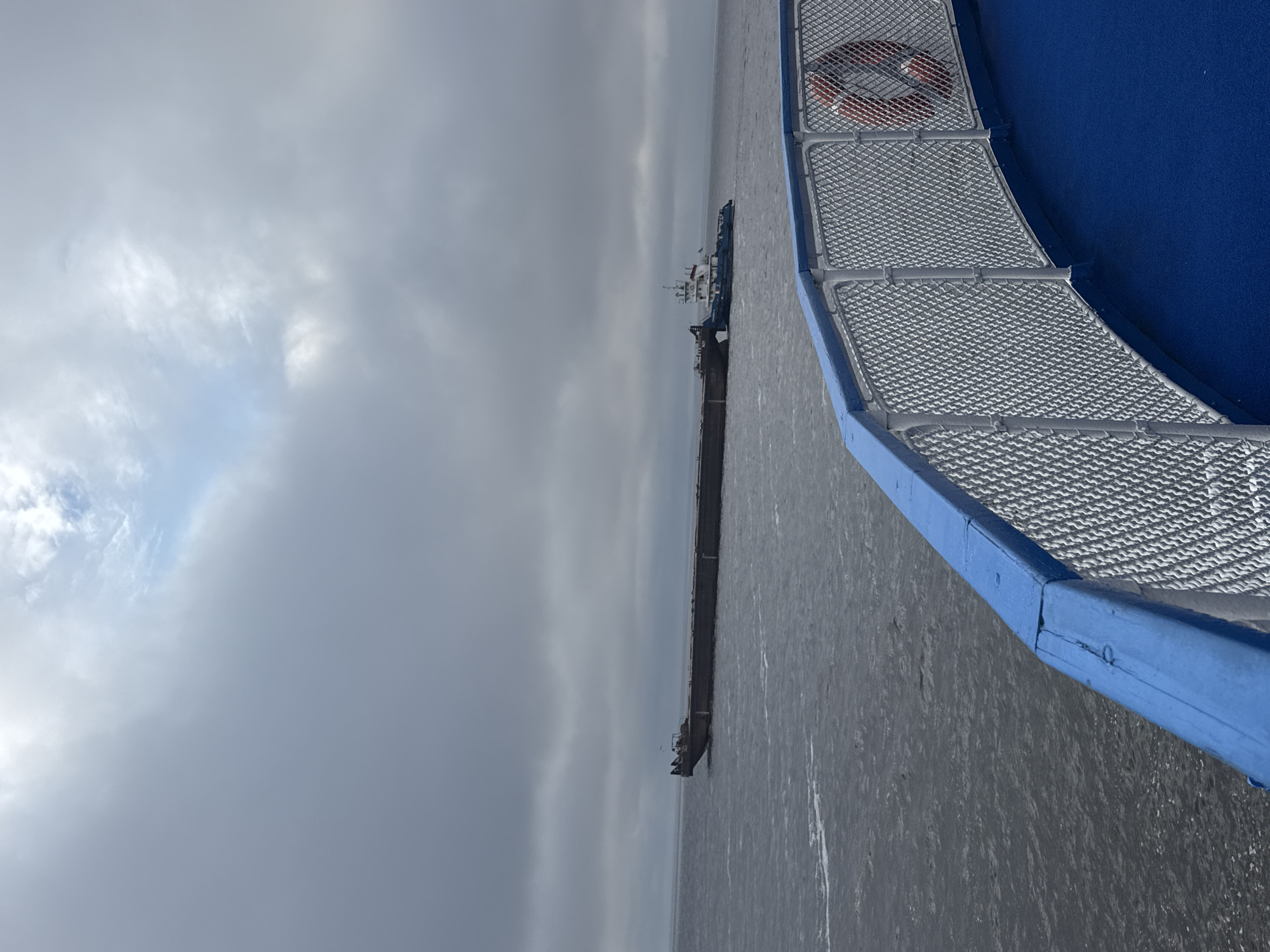 An aerial shot captures a tugboat pushing a barge laden with cargo across a wide body of water under a cloudy sky. The water is dark and choppy, suggesting a strong current or wind. The tugboat is blue and white, with a white superstructure and a red lifebuoy visible on its deck. The barge is long and flat, with a darker color and appears to be carrying bulk materials. The foreground shows the railing of a vessel, painted blue and white, with a mesh netting. A red lifebuoy is attached to the netting. The location is Laishevo, Russia.