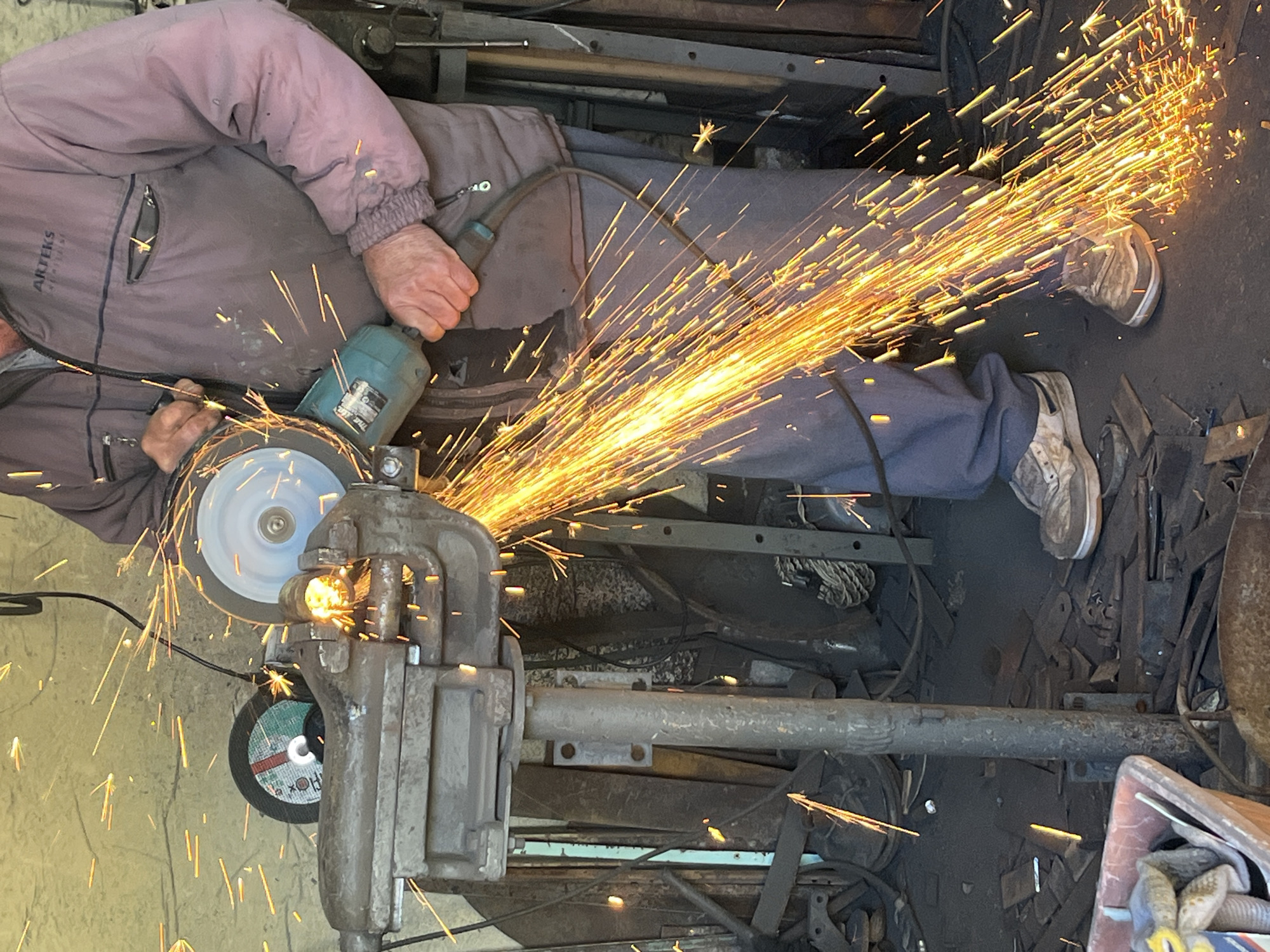 A person wearing a grey jacket and blue pants is using a grinder to cut metal held in a vise. Sparks are flying. The location is Unknown, Unknown.