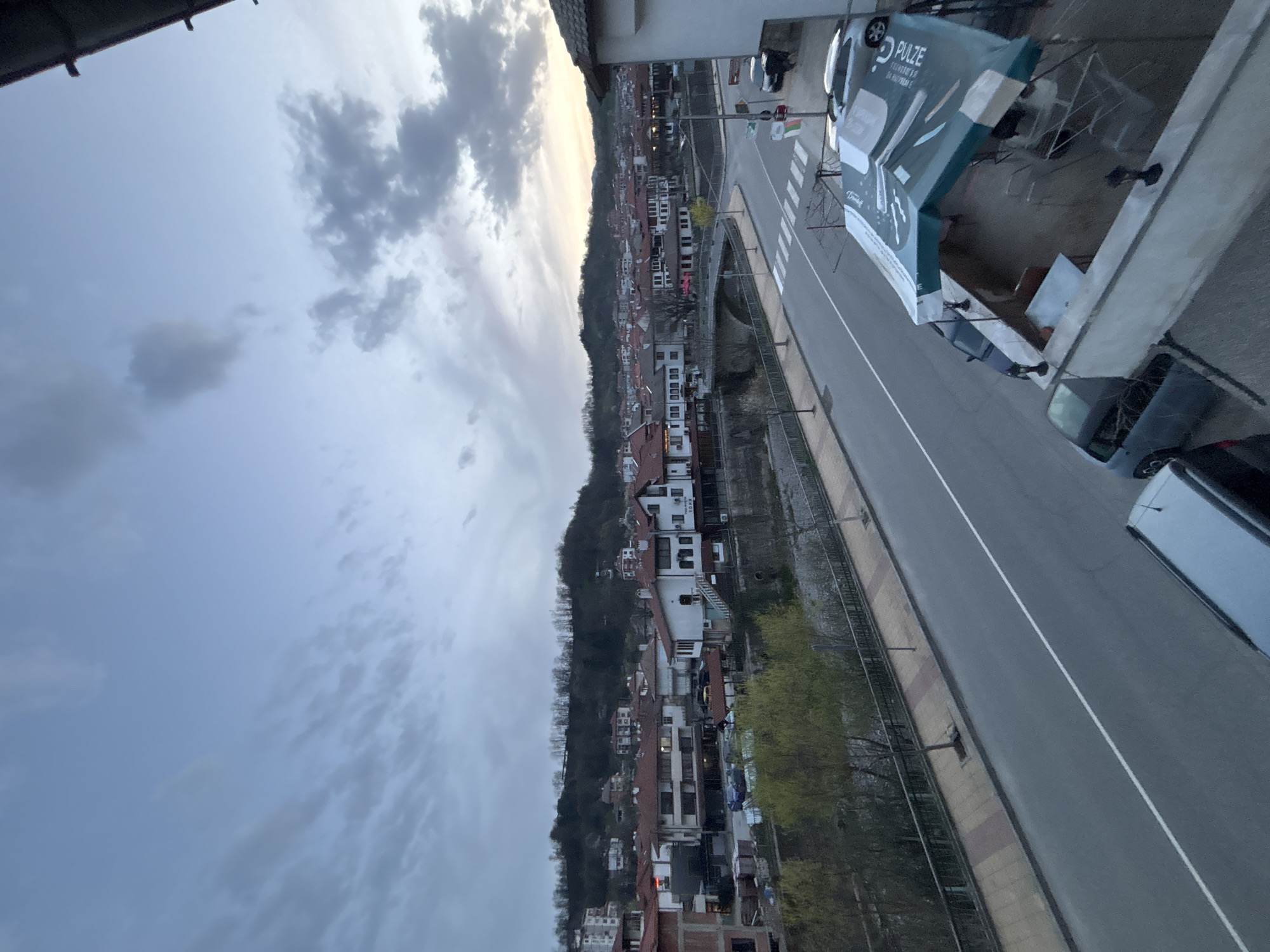 The image captures an elevated view of Zlatograd, Bulgaria, situated alongside a road that cuts through the town. On the left side of the road, the town ascends along a hillside, comprised of residential buildings with red-tiled roofs. A line of trees borders the road, and a retaining wall or a series of walls runs parallel to the road. On the right side of the road, there are parked cars and a gazebo-like structure advertising "PULZE". The sky above is overcast with cloud cover. The road appears to be mostly clear of traffic.