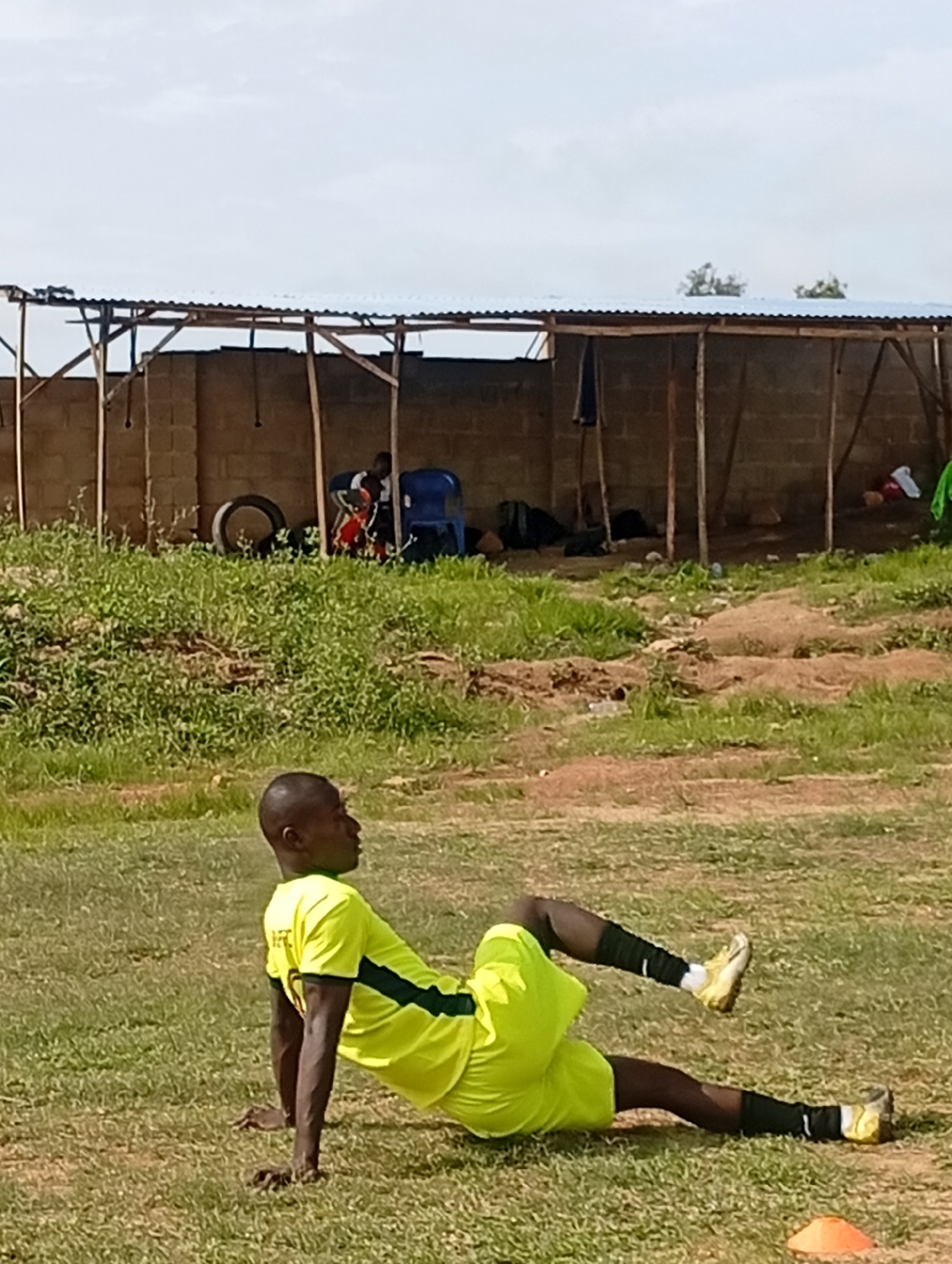 A man in a neon yellow soccer uniform is performing an exercise on a grassy field. He is on his hands and forearms, with his body lifted off the ground and his legs extended. He is wearing black socks and yellow soccer cleats. The background shows a brick wall with a corrugated metal roof structure, typical of an outdoor sports facility or training ground in Jos, Nigeria. There are some storage items like tires and blue chairs under the shelter. The sky is overcast.