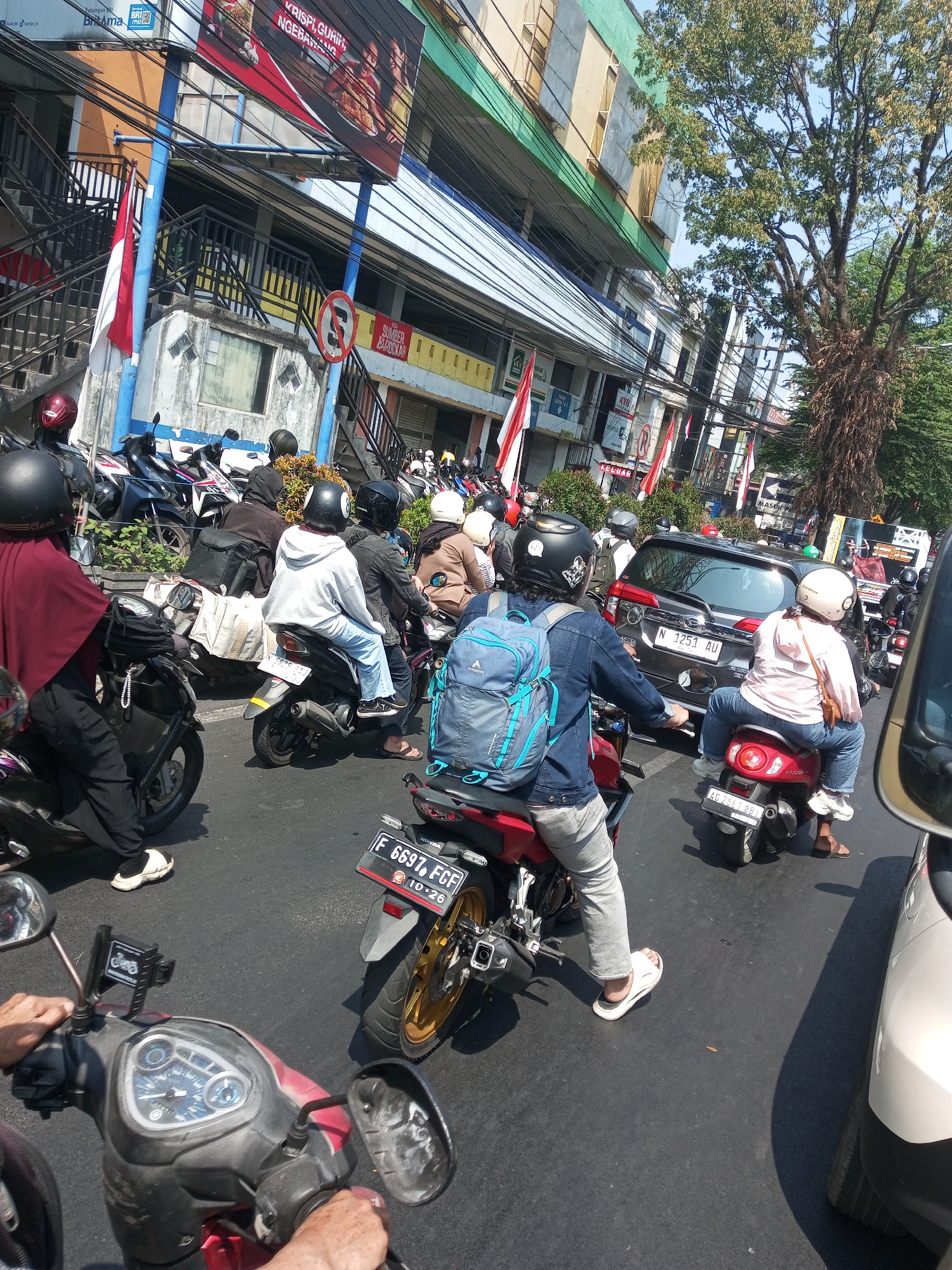This is an aerial, eye-level, medium shot of a street scene in Malang, Indonesia, featuring a traffic jam with multiple motorcycles and a car. The lighting suggests it is daytime with bright sunlight.
In the foreground, a person wearing a maroon hijab, black pants, and white shoes is riding a motorcycle. The motorcycle's handlebars and instrument panel are visible, as are the hands of the rider. To the right and slightly behind this motorcycle is another red motorcycle with a rider wearing a denim jacket and grey pants, and a passenger wearing a light pink shirt and blue jeans, both on motorcycles. The passenger is wearing a white helmet. A blue backpack is visible on the rider's back. The license plate of this motorcycle reads "AG 2367 BB1".
Further back, a dark-colored car with a license plate "N 1251 AU" is stopped in traffic. Several other motorcycles are visible around the car and in the background, some with riders wearing helmets. The environment includes buildings lining the street, with signs and flags displayed. One building has a large billboard with Indonesian text and images of food. Another building has a blue facade and stairs. A tall tree with dense foliage stands to the right of the scene. The road surface is asphalt. The presence of many motorcycles indicates a common mode of transportation in this area.