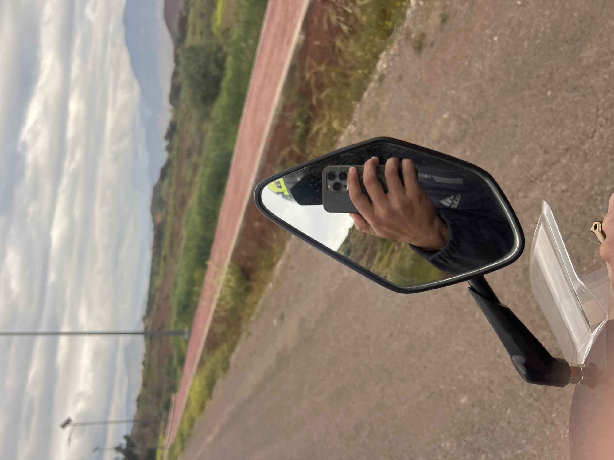 The image shows the side mirror of a motorcycle, reflecting a hand holding a smartphone to take a photo. The background features a landscape with a road, green vegetation, and mountains under a cloudy sky. The location is suggested to be in the city of Bin El Ouidane, Morocco.