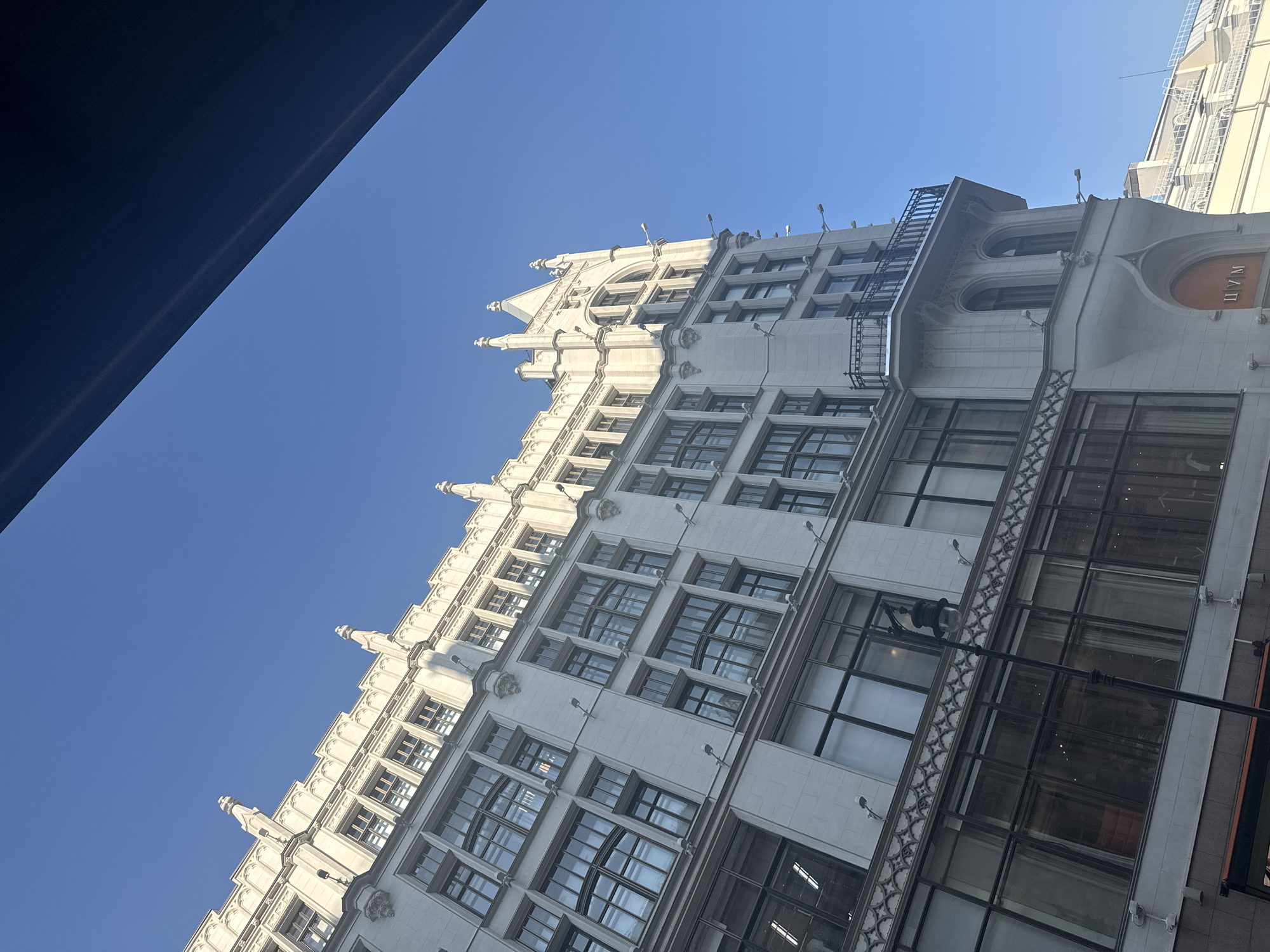 The image shows a tall, ornate white building with a clear blue sky as the background. The building has many windows, some with decorative framing. The architectural style appears to be Gothic Revival, with pointed arches, pinnacles, and intricate stonework. A balcony with a wrought-iron railing extends from one of the upper floors. On the ground level, there are large display windows. The perspective is from below, looking up at the building. A dark, angled shape obscures the top right portion of the image, suggesting the viewer is looking from inside or under something. The building appears to be a commercial establishment, possibly a department store, as indicated by the visible lettering "ГУМ" above an entrance. There are no people visible in the frame.