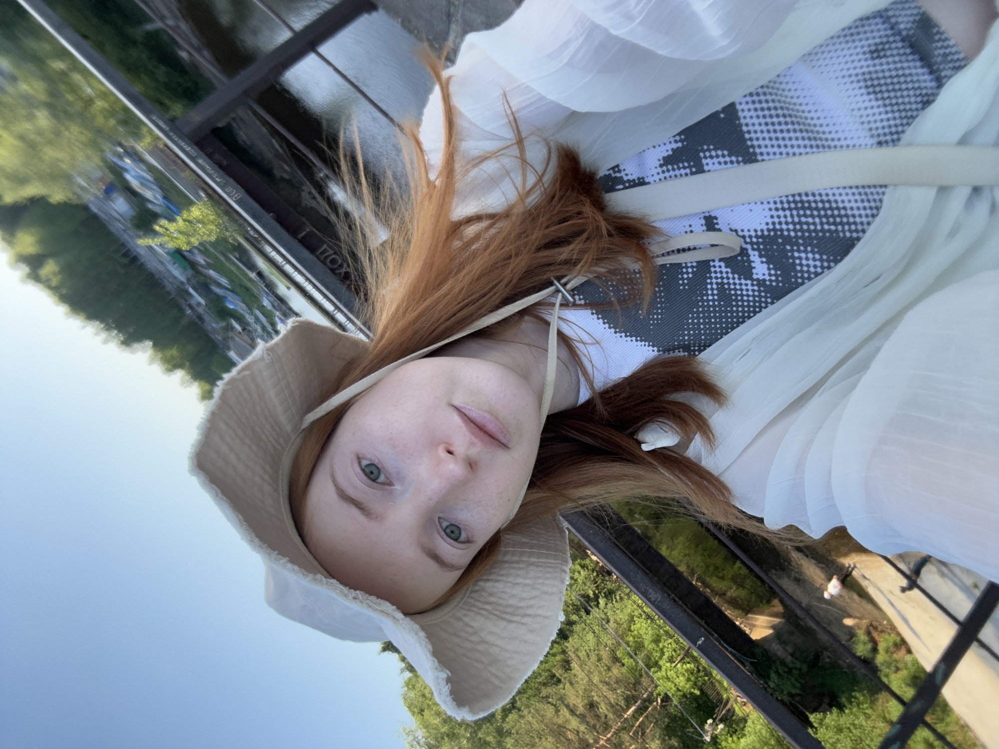 A young woman with red hair and a light-colored hat is looking at the camera. She is wearing a white top and a black and white patterned top underneath. She is standing on a bridge or walkway, with trees and water visible in the background. The location is Chaikovsky, Russia.