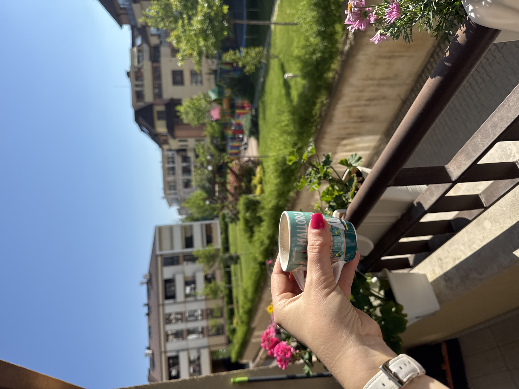 A woman's hand holds a cup with "MALLORCA" printed on it, likely on a balcony. The hand has a white watch and red nail polish. The balcony has plants in white containers. In the background, there is a green park area and several multi-story buildings under a clear blue sky, typical of an urban setting. Given the architectural style of the buildings and the overall appearance of the scene, this is likely located in Sofia, Bulgaria.