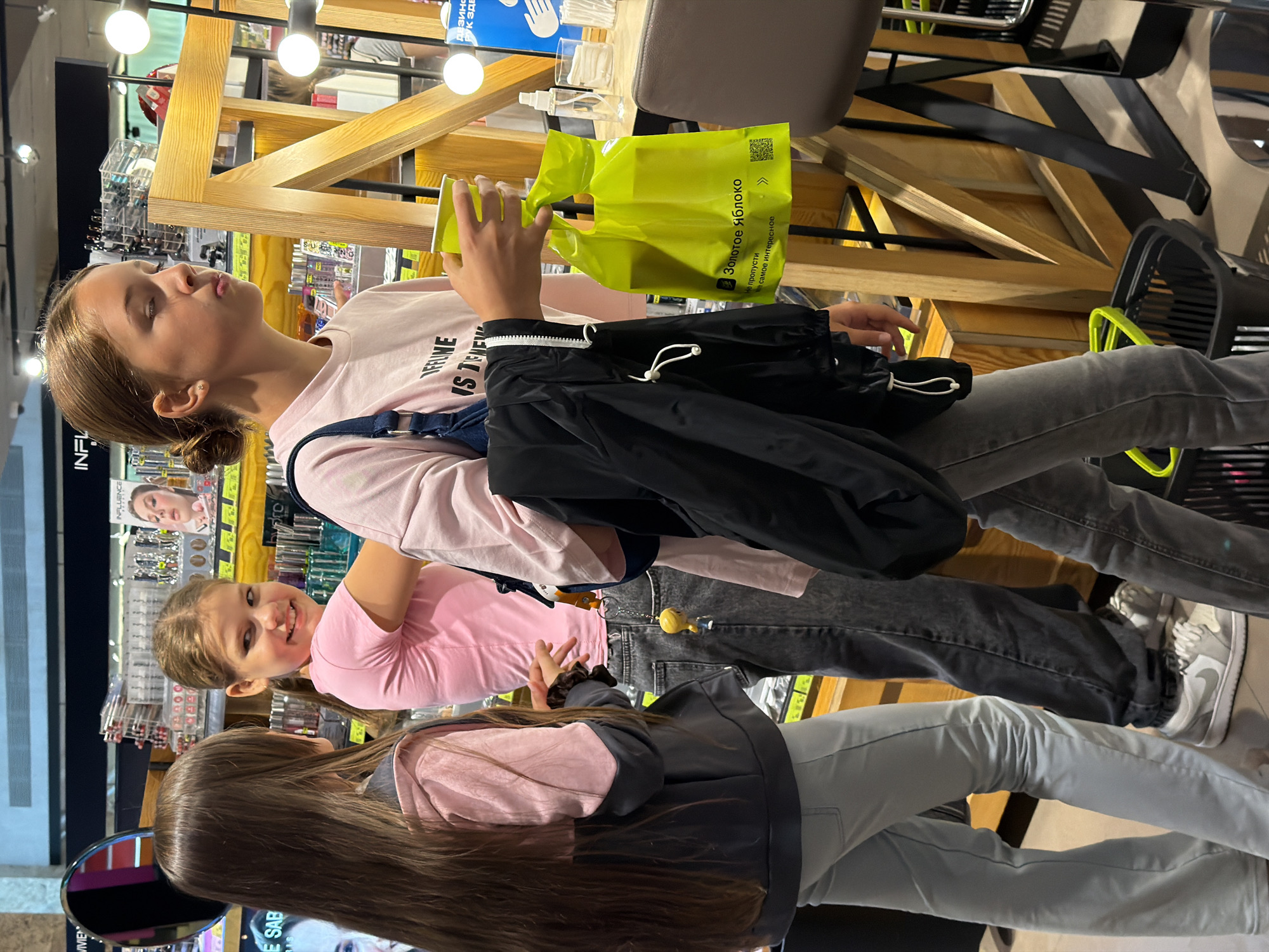 Three young girls are standing in what appears to be a cosmetics or beauty store, with shelves stocked with products in the background. The girl on the left has long, dark brown hair and is wearing a pink and grey hooded jacket over light blue pants. She is turned away from the camera, looking towards the middle girl. The middle girl has blonde hair in a braid and is wearing a pink t-shirt and dark grey jeans. She is smiling and looking over her shoulder at the girl on the left. She is holding a black jacket over her arm. The girl on the right has her hair pulled back in a bun and is wearing a pink t-shirt. She is holding a yellow cup and a bright yellow shopping bag with Russian text on it. She is looking to the right and appears to be chewing something. The environment includes wooden shelves, display stands with lights, and some retail merchandise. The lighting is bright, indicative of an indoor retail space.