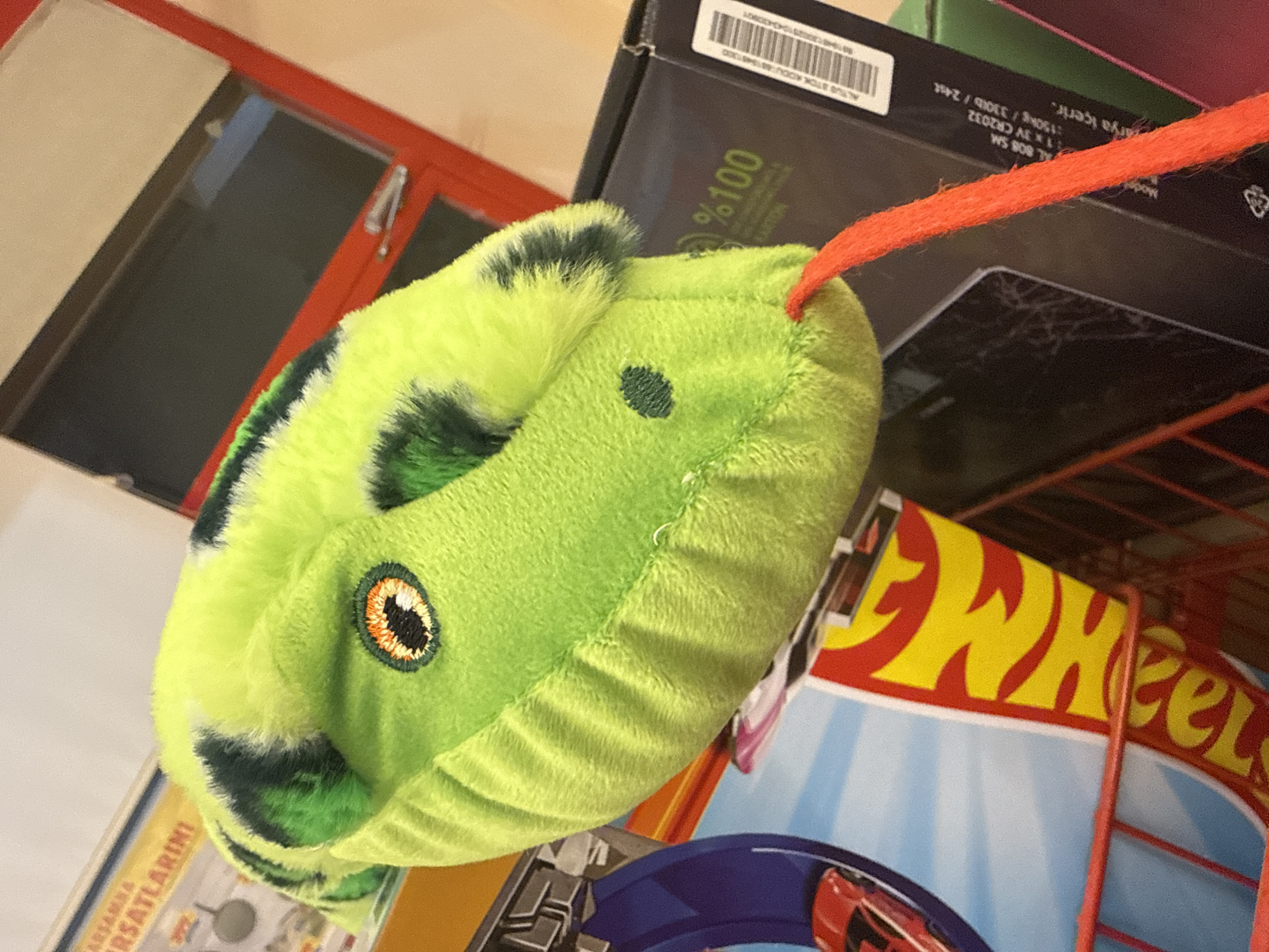 Here's a forensic description of the image:
**Subjects and Objects:** The image features a green, plush toy with a single embroidered eye and black markings, possibly representing a caterpillar or similar creature. It is attached to an orange string. Below the toy are a Hot Wheels magazine and a product box, the latter with printed details.
**Environment and Location:** The image seems to be indoors. Based on text OCR, the items may be located or related to Alanya, Turkey.
**Additional details:** The product box contains text including "ALTUS STOK KODU" and a barcode.