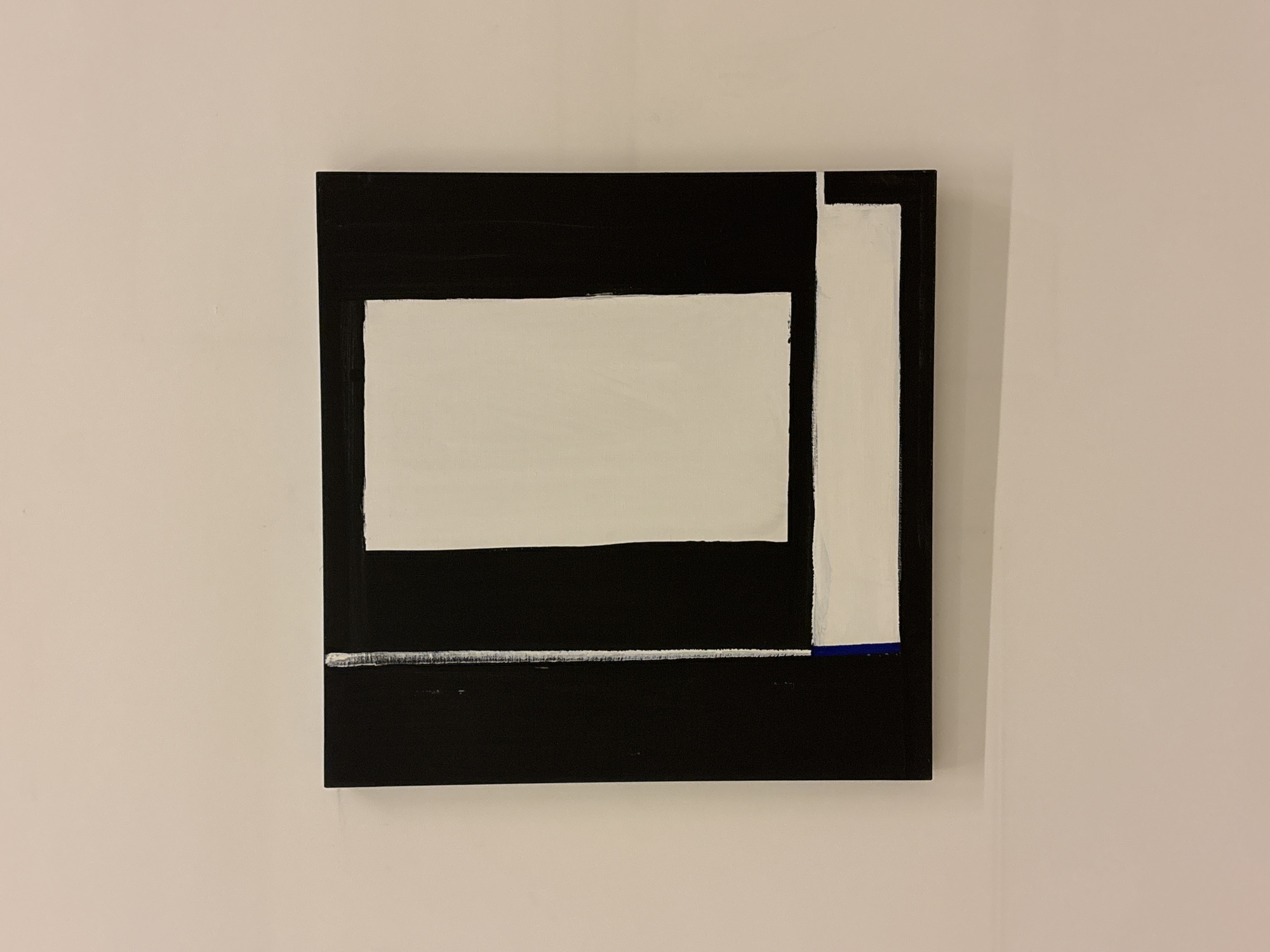 A square, abstract painting hangs on a light beige wall. The painting features a black background with a horizontal white stripe near the bottom and a white rectangle centered higher. A white stripe runs vertically along the right side. There is a small blue section at the end of the vertical stripe, where it meets the horizontal stripe. There is no discernible location in São Paulo, Brazil, depicted or implied. There are no people, subjects, actions, or interactions visible in the scene.