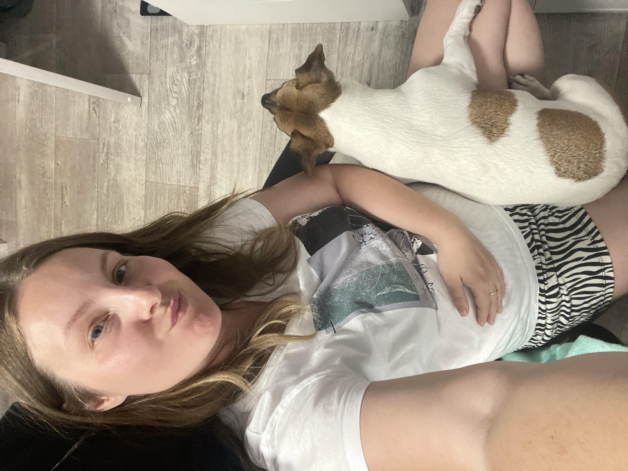 A woman lies on a surface with a Jack Russell Terrier dog lying on top of her legs. The woman is fair-skinned with light brown hair and blue eyes. She wears a white t-shirt and zebra-striped shorts. The dog is white with brown markings on its head and body. The location appears to be indoors, with light-colored wood flooring and white furniture visible in the background. The exact location is not discernible from the image.