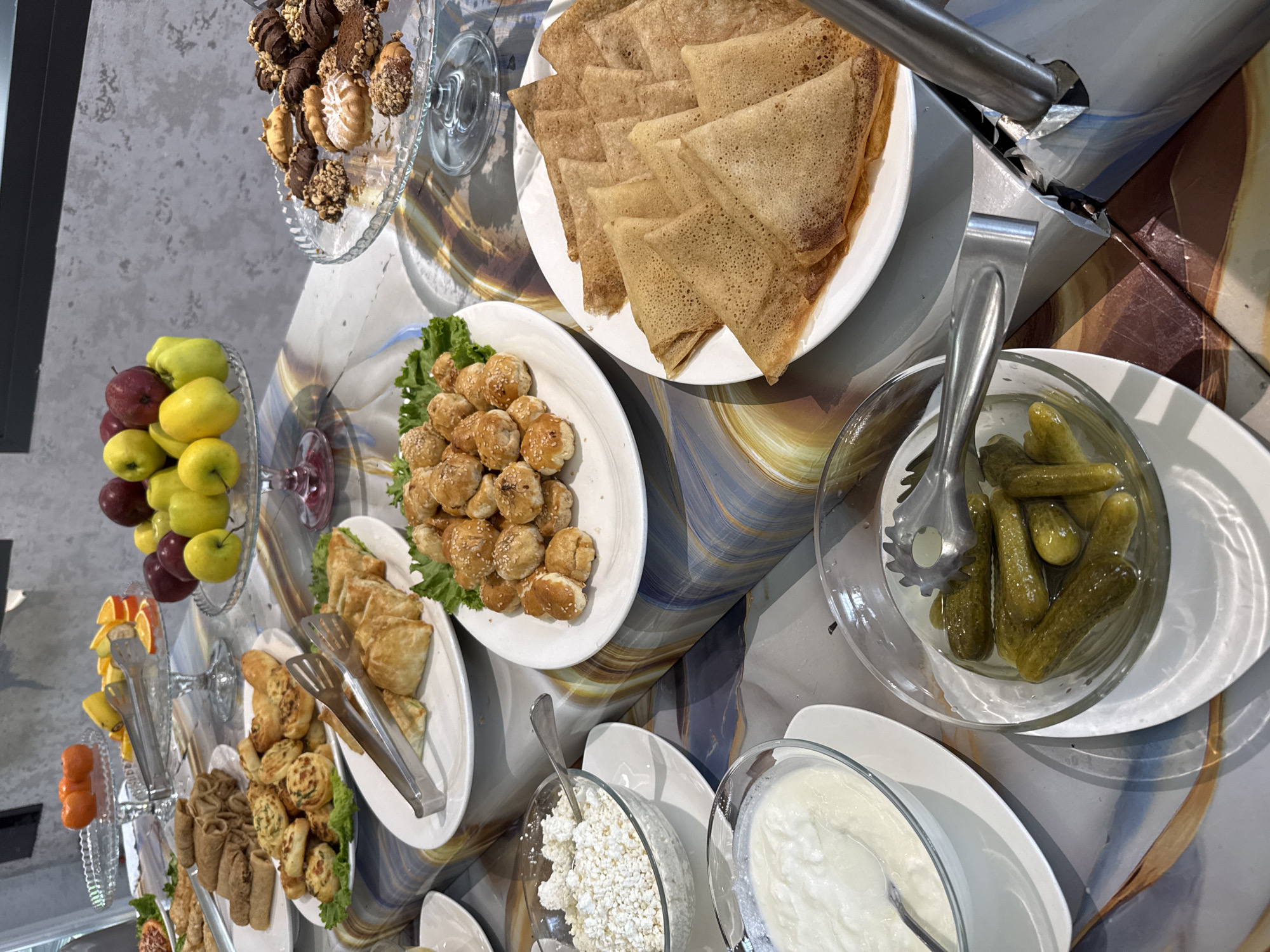 The image shows a buffet-style table with various food items. The scene includes plates of pancakes, small pastries with sesame seeds, pickled cucumbers, cottage cheese, and yogurt. There are also glass platters with apples, oranges, and assorted cookies. Serving tongs are present near the food. The table has a marble-like surface. Based on the presence of Central Asian dishes, the location is likely Tashkent, Uzbekistan.