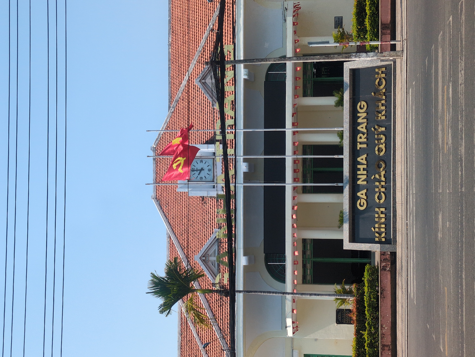 The image depicts the Nha Trang Train Station in Tây Nha Trang, Vietnam. The station building is a light-colored structure with a red tile roof. Two Vietnamese flags are visible, positioned next to a clock. A sign in Vietnamese reads "GA NHA TRANG KÍNH CHÀO QUÝ KHÁCH" (Nha Trang Station Respectfully Welcomes Guests). A row of electric wires is visible against a clear blue sky. A palm tree is present in the foreground. The station is adjacent to a road.