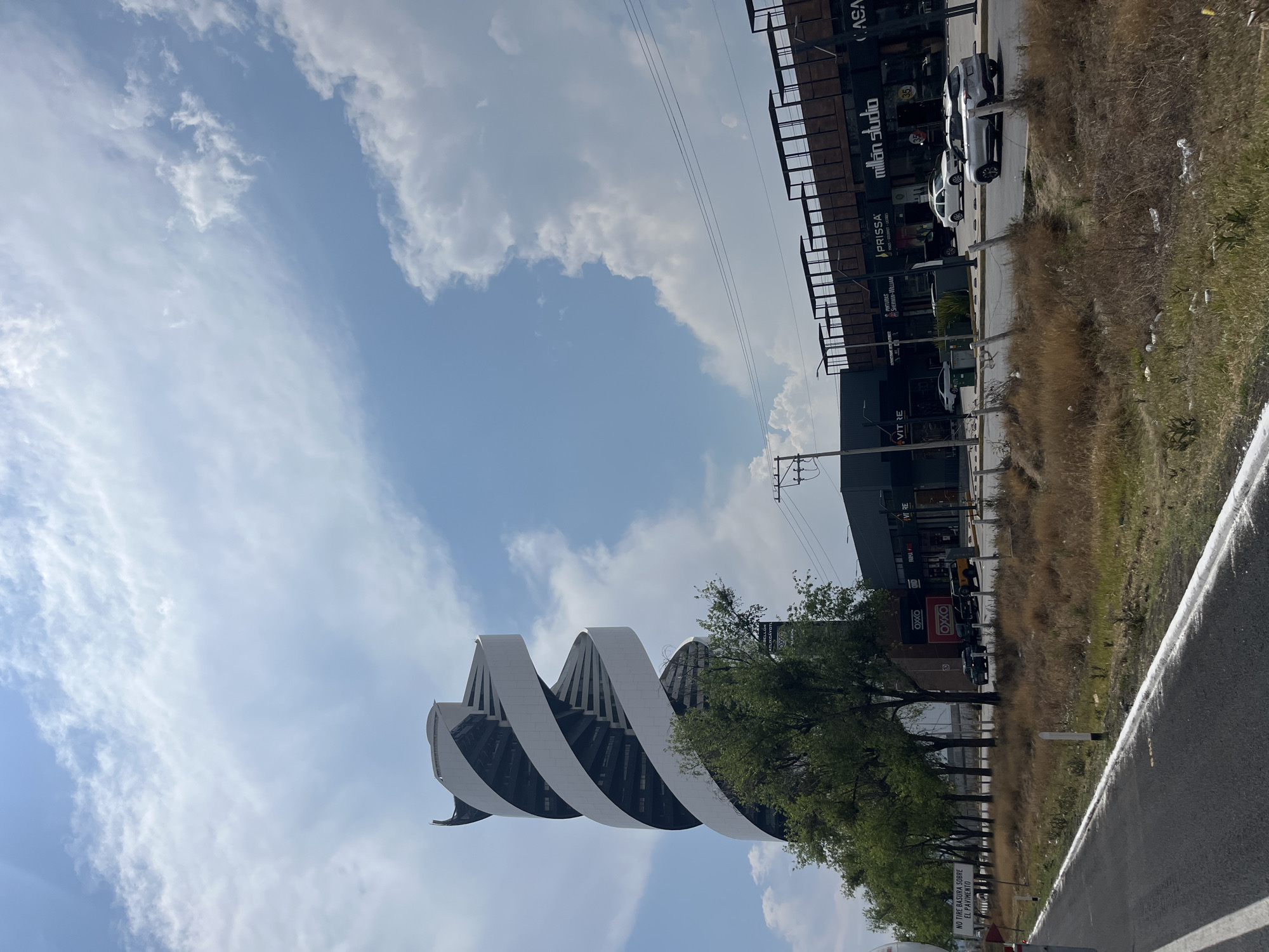 The image depicts a view of a distinctive architectural structure in San Bernardino Tlaxcalancingo, Mexico. The building is characterized by its spiraling, white and black design, rising against a backdrop of a partly cloudy sky. Adjacent to the building, there's a line of trees providing green contrast, and a commercial area is visible, with storefronts including "millan studio", and "PRISSA". A roadside with dry grass and litter leads to a street with parked cars. Street signs, including one reading "NO TIRE BASURA SOBRE EL PAVIMENTO", can be seen. The image is shot from a low angle and has been rotated.