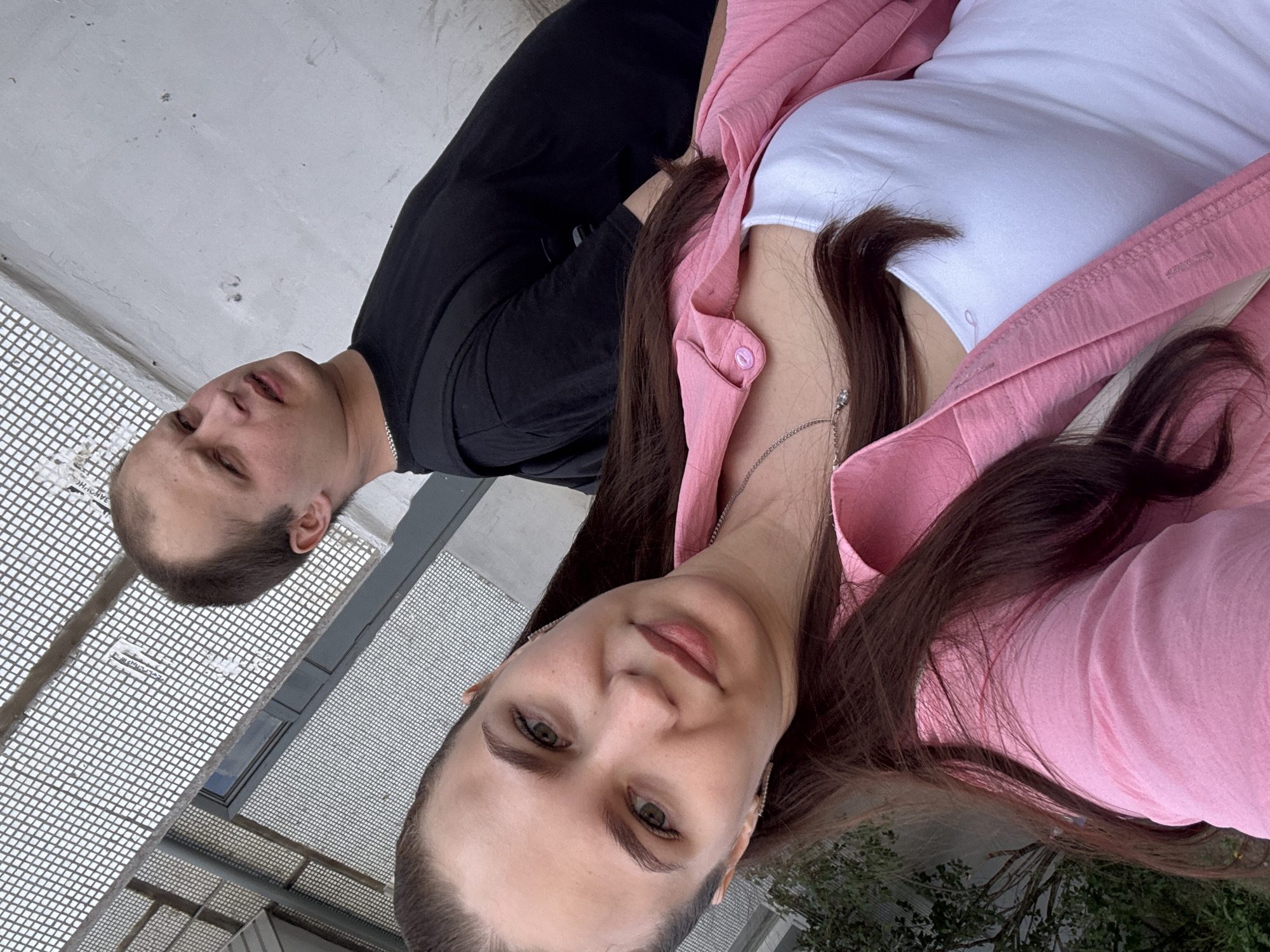 A selfie of two people on a roof. One is a man with short brown hair wearing a black t-shirt. The other is a woman with long brown hair wearing a white t-shirt and a pink button-up shirt. The location is likely in the city of Chaikovsky, Russia. The background shows a metal grate and a building.