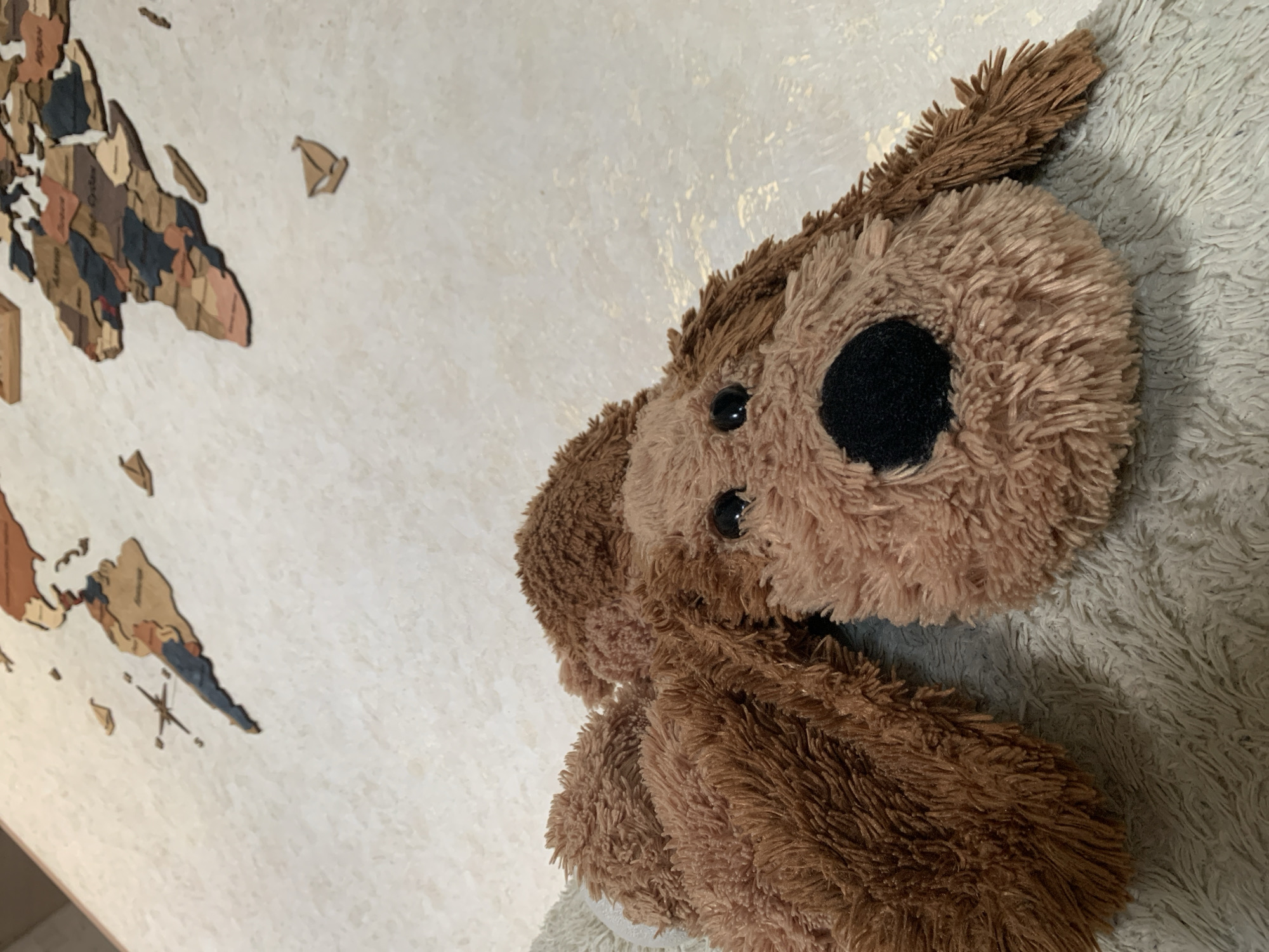 The image displays an indoor scene featuring a brown plush dog toy and a decorative world map on a wall. In the foreground, a brown, shaggy plush dog toy with a black nose and black eyes rests on a soft, light-colored, fluffy surface, likely a blanket or rug. The texture of the toy's fur is prominent, showing various shades of brown.
Behind the toy, a large wall decoration depicting a world map is visible. This map appears to be crafted from multiple pieces of wood or similar material, featuring different colors such as brown, beige, and dark grey/blue for the landmasses. Scattered around the main continents are smaller decorative elements, including what looks like a tiny wooden sailboat and a compass rose. The wall itself is light-toned with a slightly textured finish.
No activity is taking place; the scene is static. The lighting suggests an indoor setting. No people are visible, and there is no discernible text within the image. While the location context is provided as Severodvinsk, Russia, there are no direct visual cues in the image to confirm this specific location.