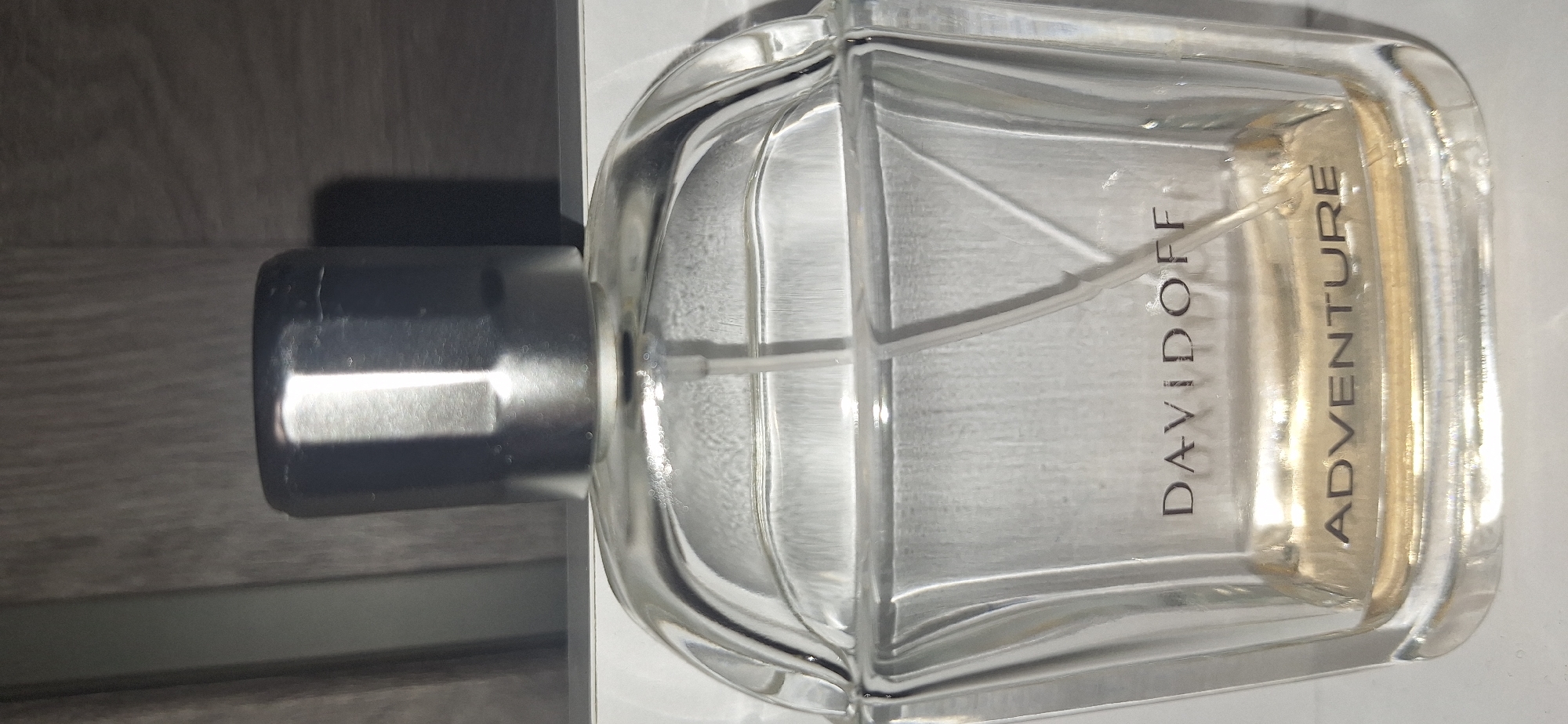 A close-up, waist-up shot shows the top portion of a clear glass perfume bottle with a silver cap. The bottle contains a golden-colored liquid. The brand name "DAVIDOFF" is printed in black capital letters on the upper portion of the bottle, and below it, the word "ADVENTURE" is also printed in black capital letters. The bottle is placed on a white surface, and a blurred background of vertical wooden panels is visible behind it. The lighting is somewhat harsh, creating reflections and highlights on the glass and silver cap. The image does not provide any information about the location in Bila Tserkva, Ukraine.