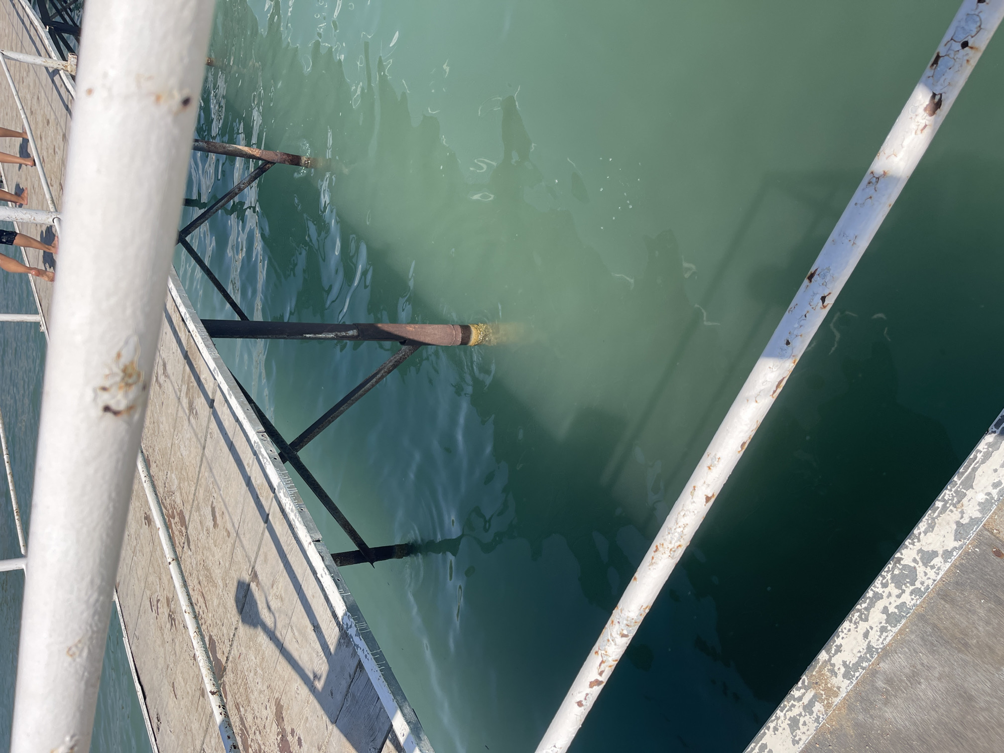 The image depicts a section of a pier extending over a body of turquoise water. The pier is constructed from wooden planks and supported by dark metal beams and poles, some of which are submerged in the water. White railings, showing signs of rust and peeling paint, line the edges of the pier. The water is clear enough to see the submerged parts of the pier's structure. The surface of the water is slightly rippled, reflecting the sunlight. In the upper part of the image, the legs of at least three individuals are visible, standing on the pier. They are dressed in casual attire, with one person wearing black shorts. The overall scene suggests a warm, sunny day at a waterfront location. The image does not provide information to confirm its location in Ornek, Kyrgyzstan.