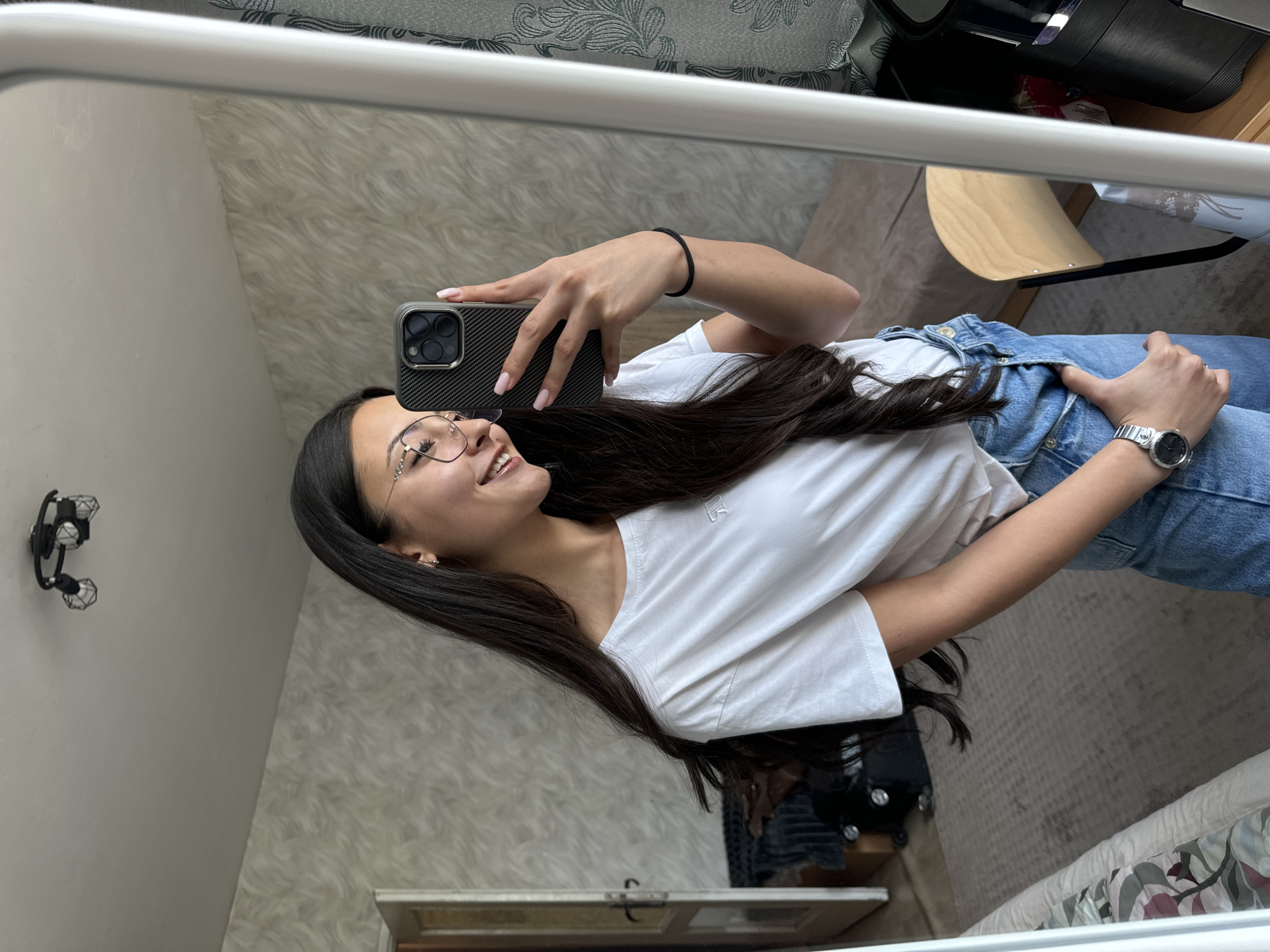 A young woman is captured in a mirror selfie, smiling broadly and looking towards the upper right of the frame, past her phone. She holds a smartphone with a dark, carbon fiber patterned case in her right hand, her light pink manicured nails visible. She has long, dark hair flowing over her shoulders and down her back. She wears clear-rimmed glasses with subtle embellishments, a plain white short-sleeved t-shirt, and light wash blue jeans. A silver-colored watch with a round face is on her left wrist, and a black braided hair tie is on her right wrist.
The setting is an indoor space, reflected in a mirror with a white frame. The background behind the woman in the reflection includes a light-colored wall with a subtle textured pattern, possibly wallpaper, and another wall with a gray and white floral or leafy patterned wallpaper. A black wall-mounted light fixture with three small cage-like lamps is visible on the textured wall. Also reflected are parts of light wooden furniture, possibly a chair, and patterned fabric that appears to be bedding or a cushion. The lighting suggests it is daytime. The scene conveys a cheerful and casual mood. No visible text is present on any objects or surfaces. The location context is Sofia, Bulgaria.