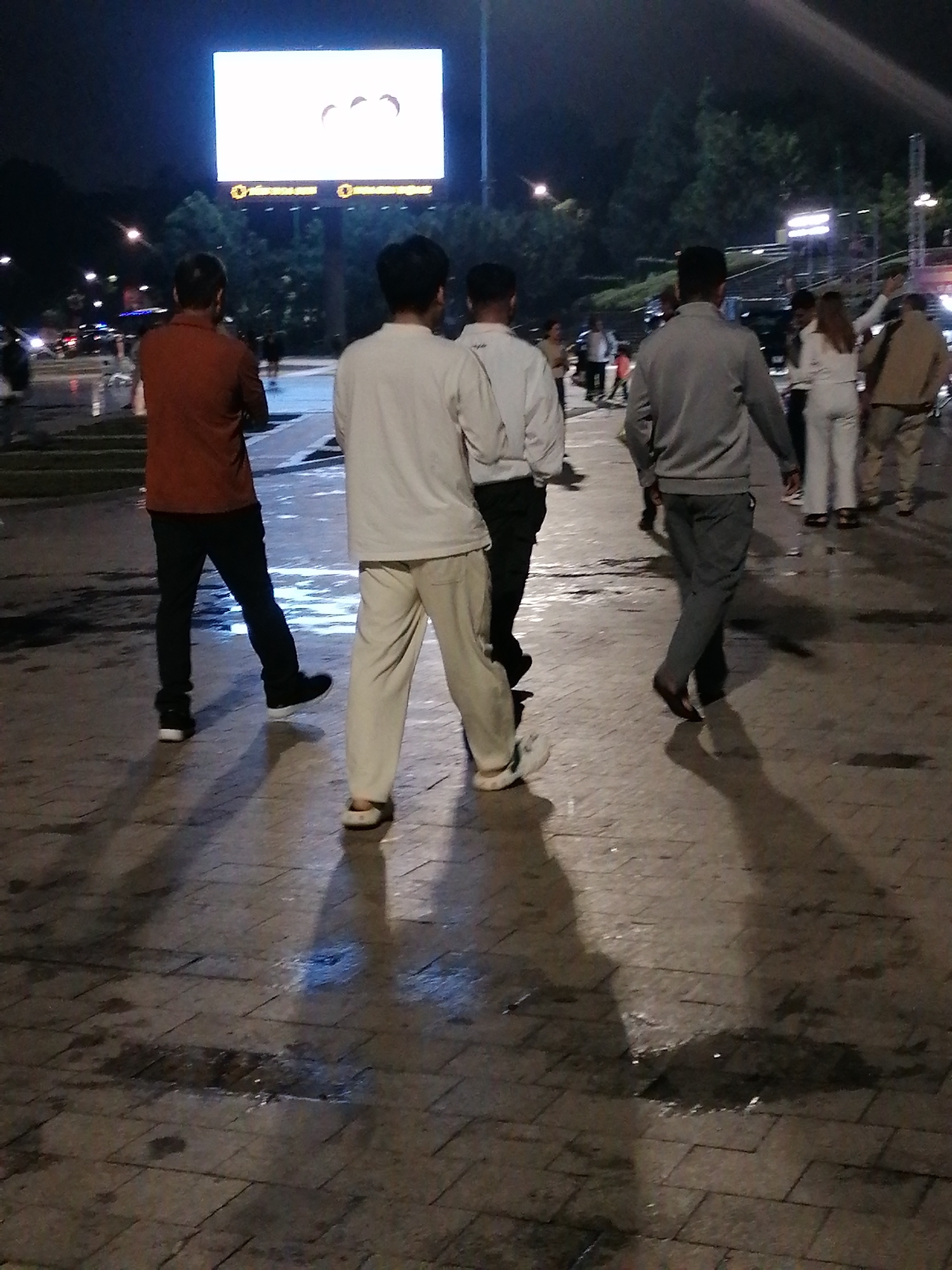 The image shows a nighttime scene on a paved walkway, possibly in Da Lat, Vietnam, given the distinct cobblestone-like paving pattern and lush, dark foliage visible in the background, characteristic of the city's cooler climate and vegetation. A large, brightly lit digital billboard is prominent in the upper left background, displaying a simple graphic of three silhouetted heads. Several individuals are walking away from the viewer, casting long shadows on the wet pavement.
The person closest to the foreground on the left wears a reddish-brown long-sleeved shirt and dark pants, with black sneakers. To their right, a person in a white long-sleeved shirt and light-colored pants walks away. Further to the right, another individual is dressed in a grey tracksuit. In the mid-ground, several other figures are visible, including a person in white pants and a light-colored top, and another in a tan jacket. The wet surface of the walkway suggests recent rain or mist, common in Da Lat. The overall atmosphere is that of a public space at night, with artificial lighting from the billboard and streetlights creating distinct shadows.