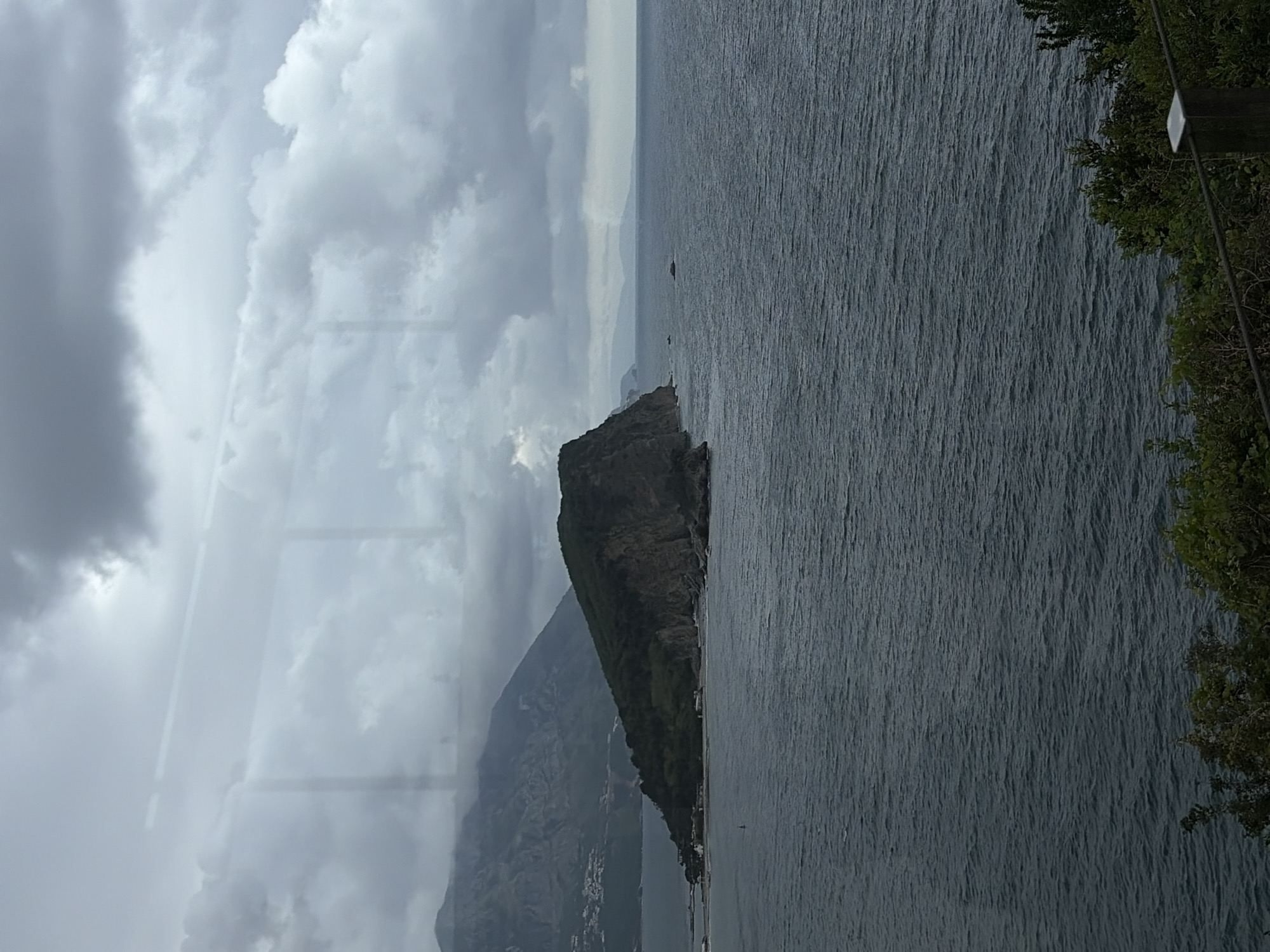 The image shows a view from a high vantage point, possibly a building or a hillside, overlooking the Adriatic Sea near Budva, Montenegro. The foreground features a body of water with a rugged coastline on the right, marked by dark rock formations and some greenery. In the middle ground, there is a land mass that leads to a distant coastline. The background is dominated by a cloudy, overcast sky, with varying shades of gray. The overall impression is one of a dramatic coastal landscape under gray weather conditions.