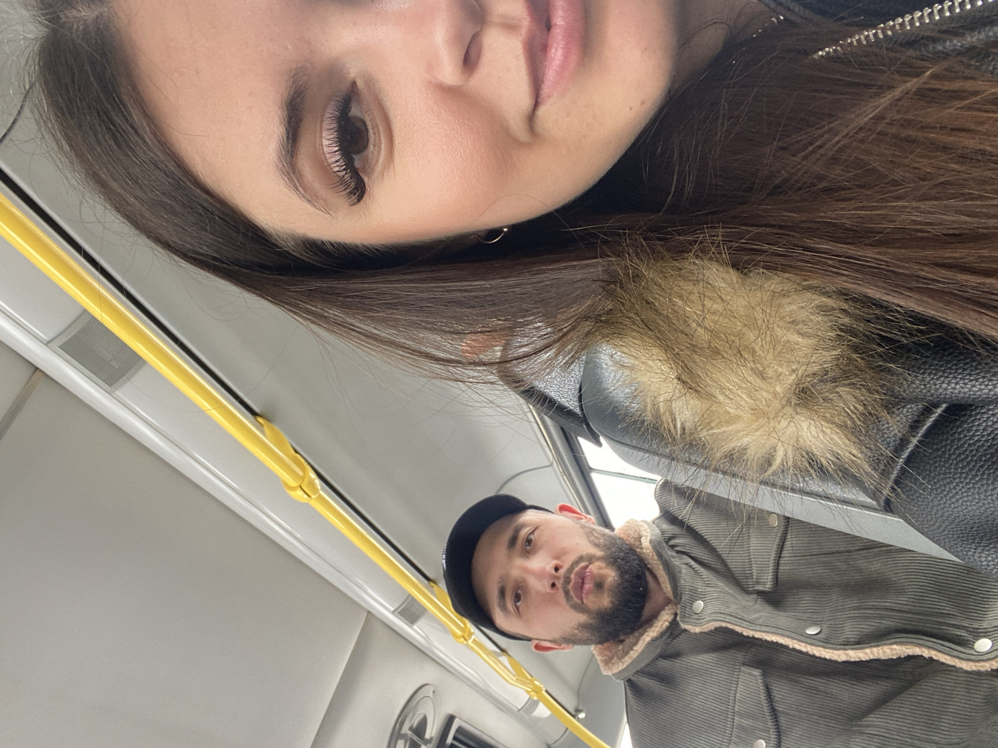Two people are visible in the image, seemingly taking a selfie. The person in the foreground has long brown hair, styled eyelashes, and is wearing a black leather jacket with a fur collar. Behind and below is a bearded man in a corduroy jacket with a shearling collar and a black baseball cap. They are both inside what appears to be a public transportation vehicle, indicated by the yellow handrail and interior fittings of a bus or tram. The background is blurred, but the interior suggests they are within the confines of an urban transport setting, likely in the city of Chaikovsky, Russia.