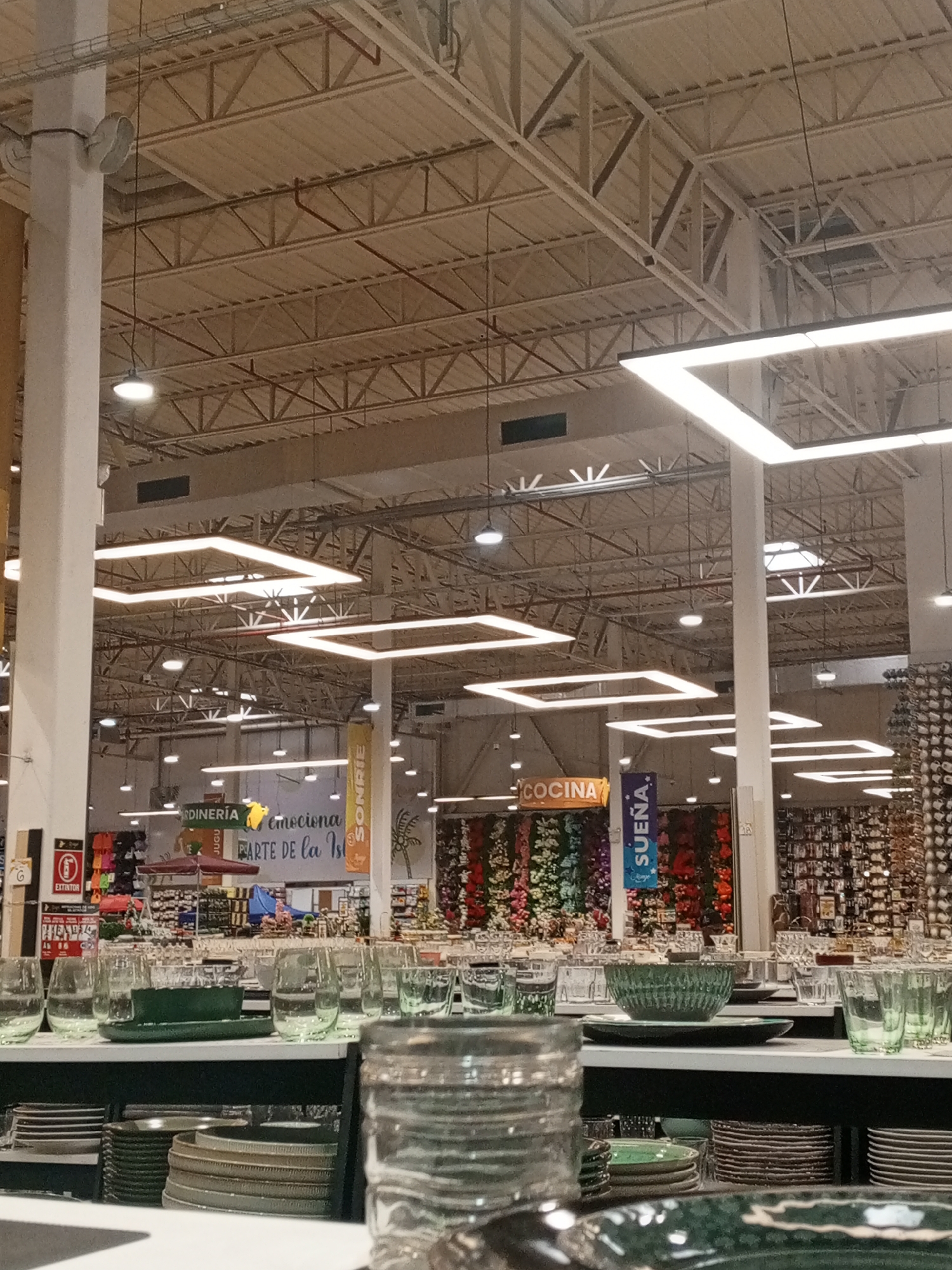 The image shows the interior of a large retail store, possibly a department store or home goods store, located in Pampatar, Venezuela. The foreground features a display of glassware and dishware in shades of green and clear glass arranged on shelves. The background shows various departments with signage in Spanish, including "JARDINERÍA," "COCINA," and "SUEÑA" (Gardening, Kitchen, and Dream). There is no people. Overhead, the store's ceiling is visible, with exposed metal beams and rectangular lighting fixtures.