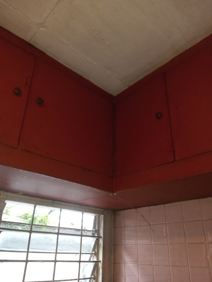 A corner of a room features old red wooden cabinets under a white ceiling. One cabinet has two doors, the other has one. The wall beneath the right cabinet is covered in small pink tiles. A window with a metal frame is visible under the left cabinet, offering a glimpse of greenery outside. A gap exists between the cabinets, revealing the wall behind.